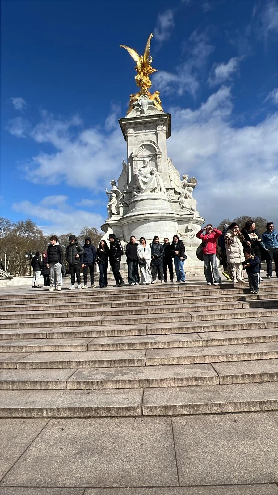 Group of people standing on steps in front of a large monument with sculptures and a golden winged figure at the top, under a partly cloudy blue sky.