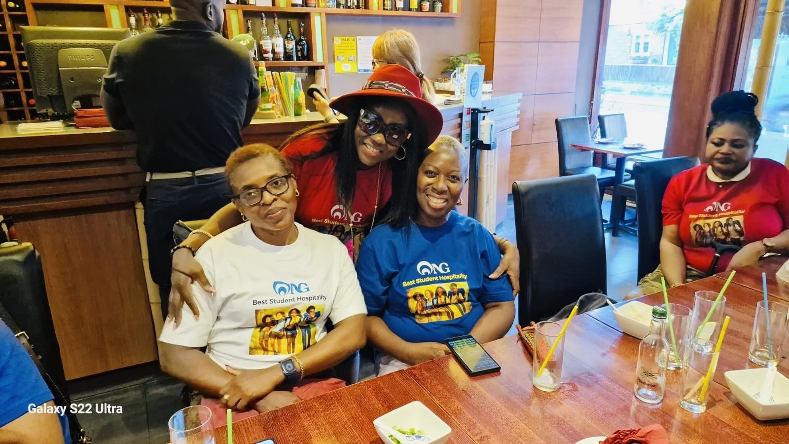 Four women sitting at a restaurant table, three wearing shirts with 'NG' and 'Best Student Hospitality' logos, smiling. One woman is standing behind them wearing sunglasses and a red hat, hugging two of the seated women. The table has glasses with ye