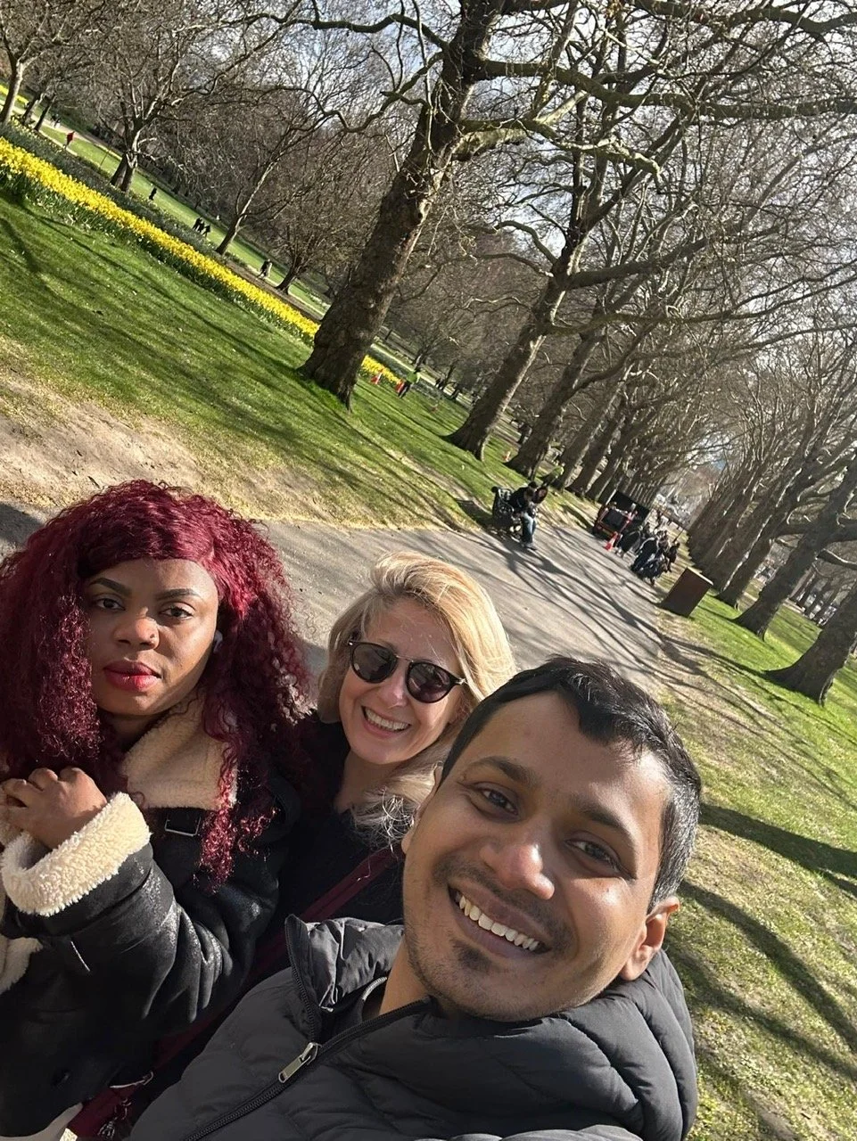 Three people smiling and taking a selfie in a park with tall trees and a pathway, some yellow flowers, and people in the background.