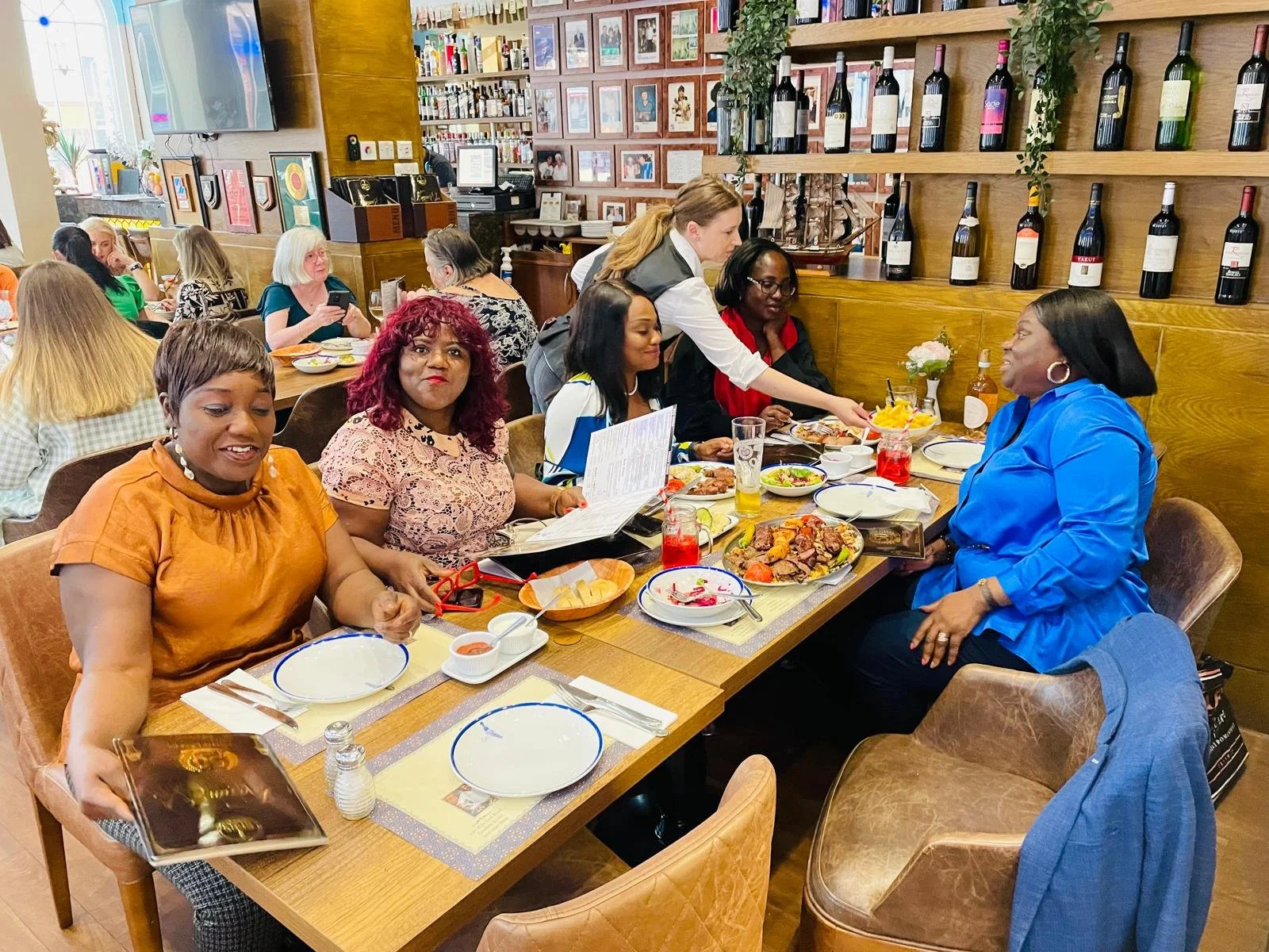 Women of diverse backgrounds enjoying a meal together at a restaurant table with various dishes and drinks, surrounded by other diners in a cozy, well-decorated restaurant.