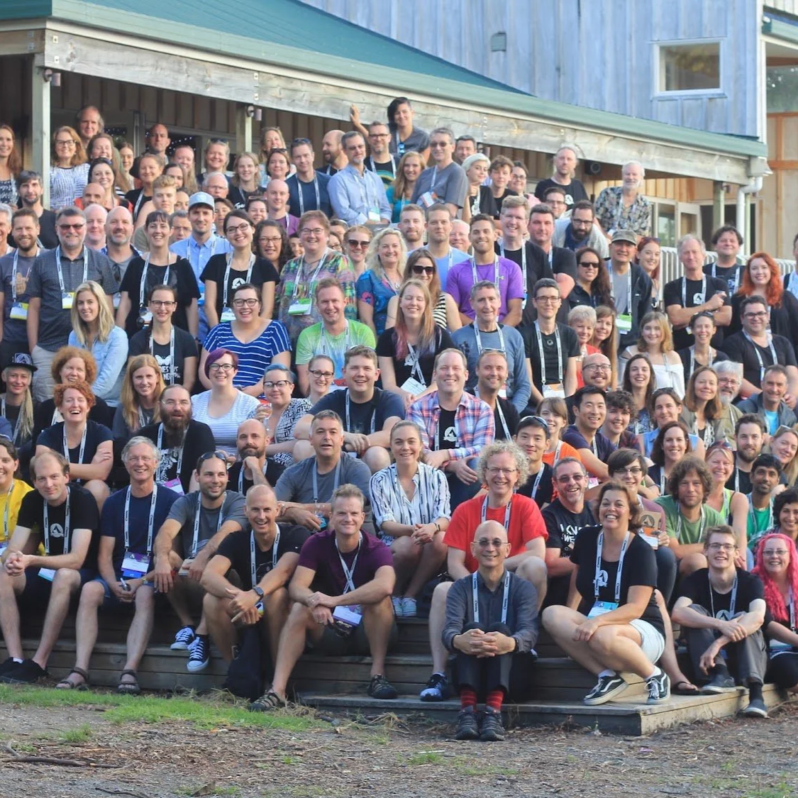 Large group of diverse people gathering outdoors on steps in front of a building, smiling and posing for a group photo at a the KiwiFoo non-conference event.