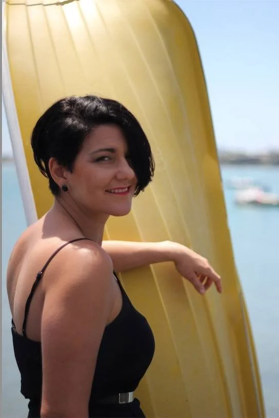 A woman with short black hair and a black dress smiling next to a yellow kayak near a body of water.