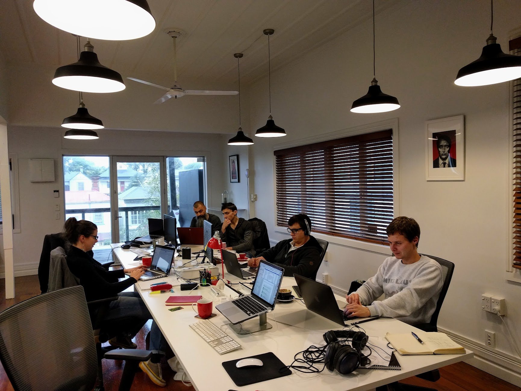 A group of six people working at a long white conference table in a bright office. They are using laptops, and some have headphones. The room has large windows with wooden blinds, ceiling lights, and framed artwork on the walls.