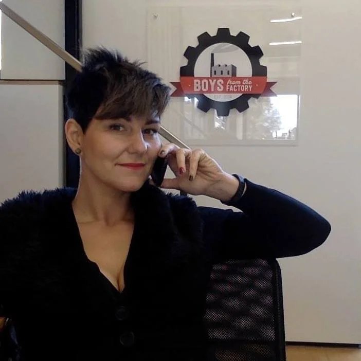 Kati Ohens talking on a cellphone inside an office with a 'Boys from the Factory' logo on the wall behind her.