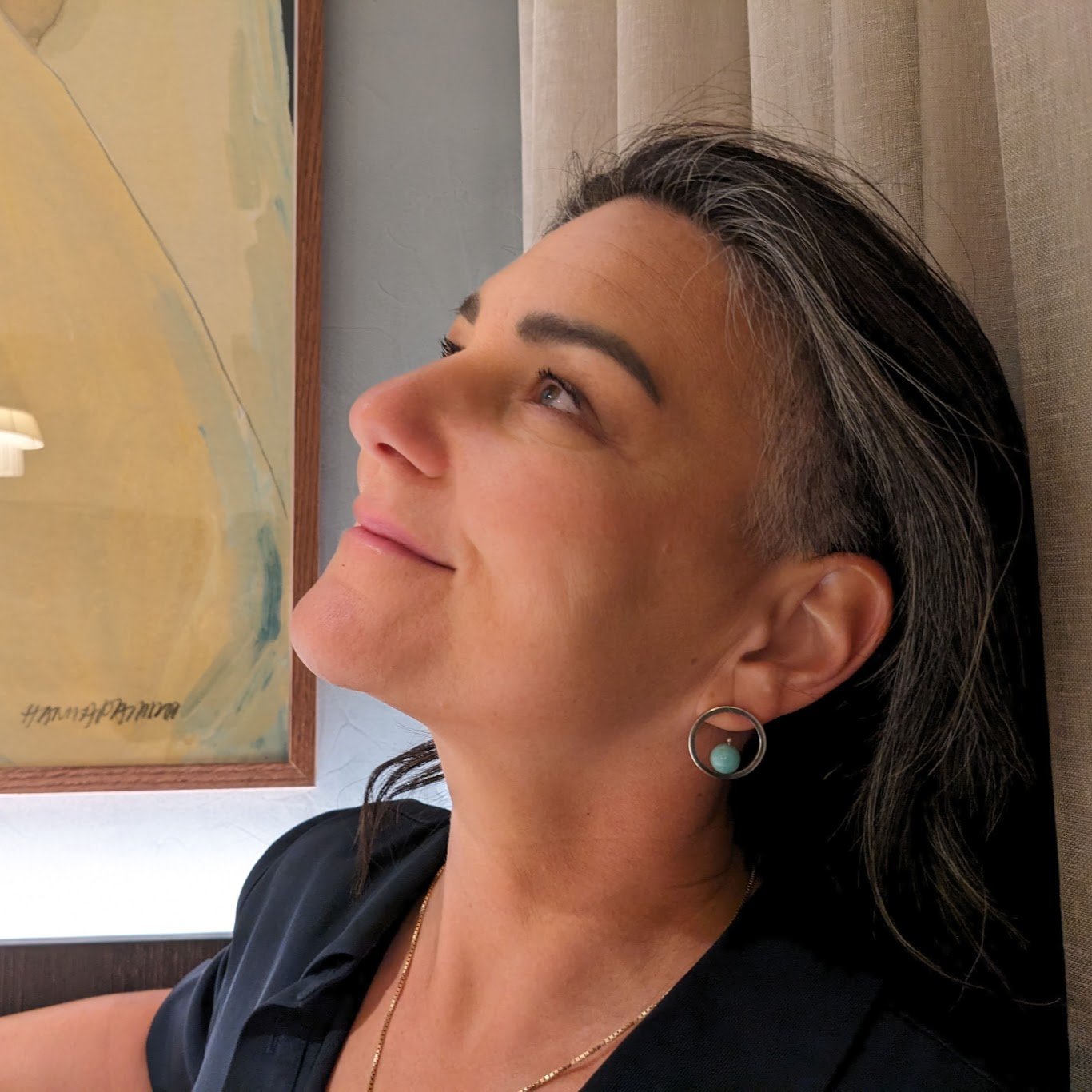 A woman with dark hair wearing a black top and turquoise earrings resting her head on a beige cushioned surface, looking upward against a background of a painted wall.