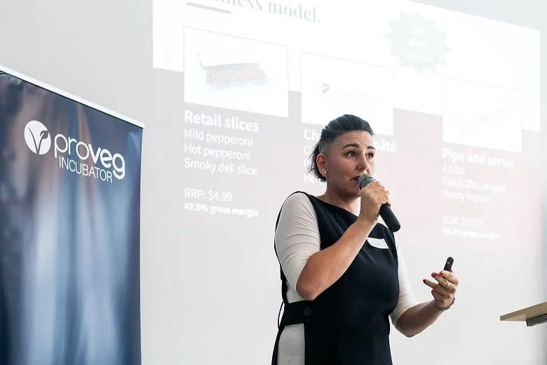 A woman speaking into a microphone during a presentation at a Provég INCUBATOR event, with a projected slide behind her.