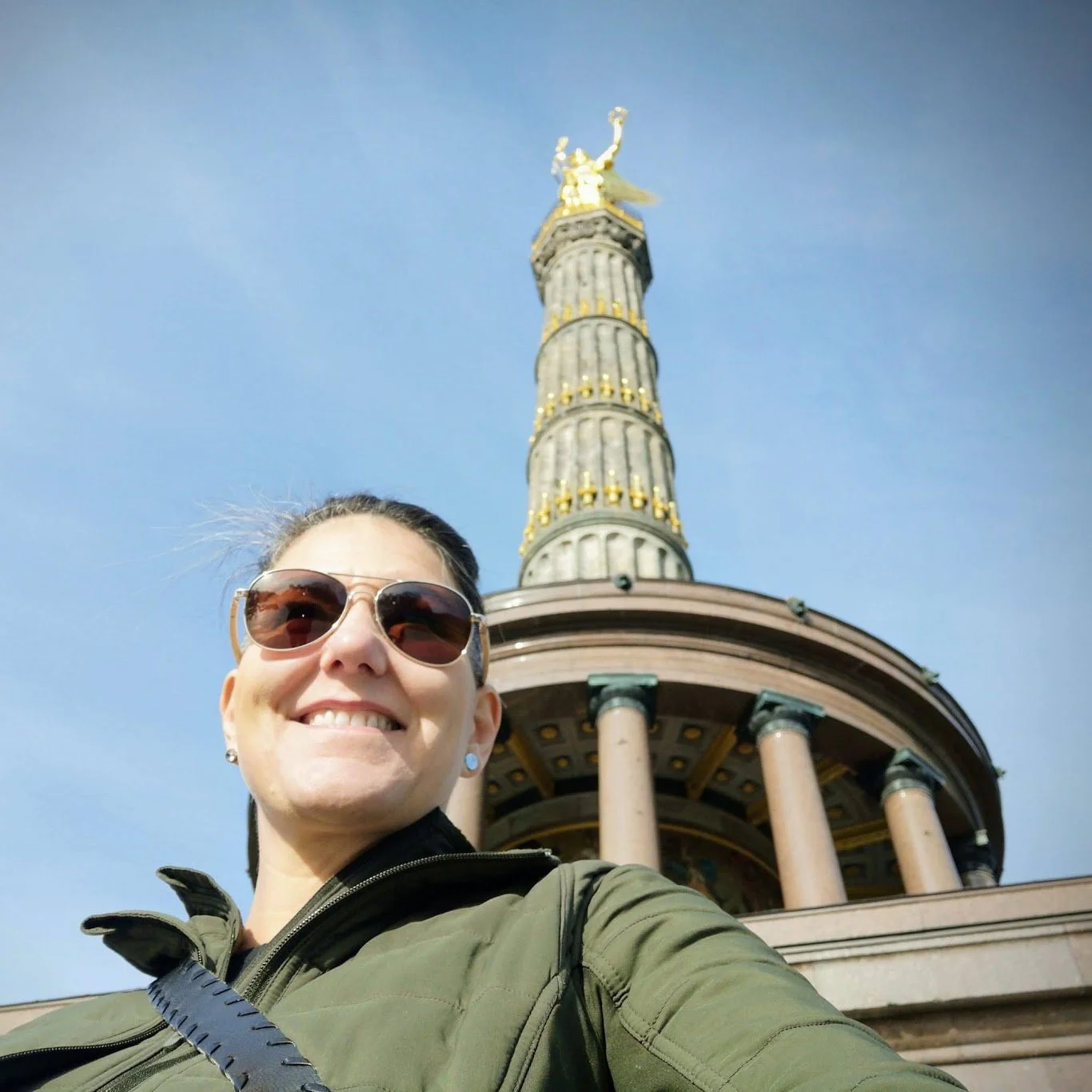 A woman smiling in sunglasses and a green jacket taking a selfie in front of the Victory Column in Berlin, Germany, with a clear blue sky in the background.