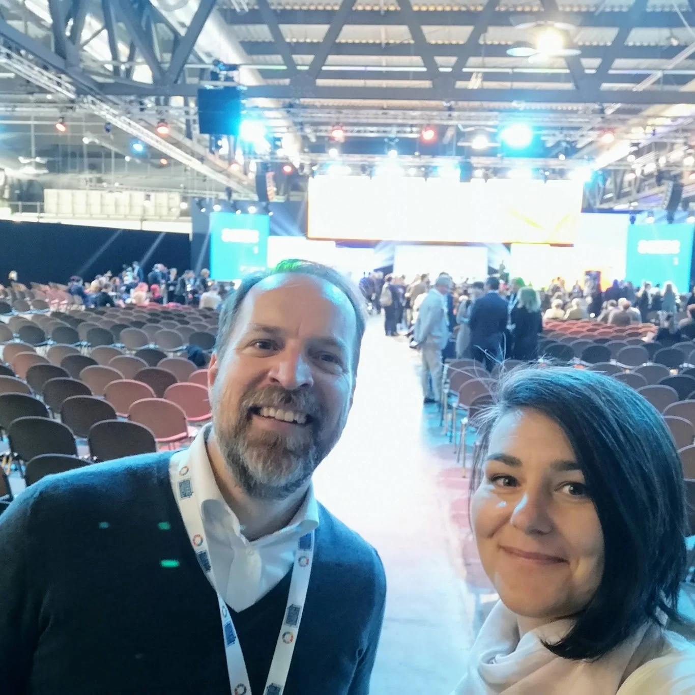 Smiling man and woman taking a selfie at a large conference or event hall with a stage and many empty chairs in the background.