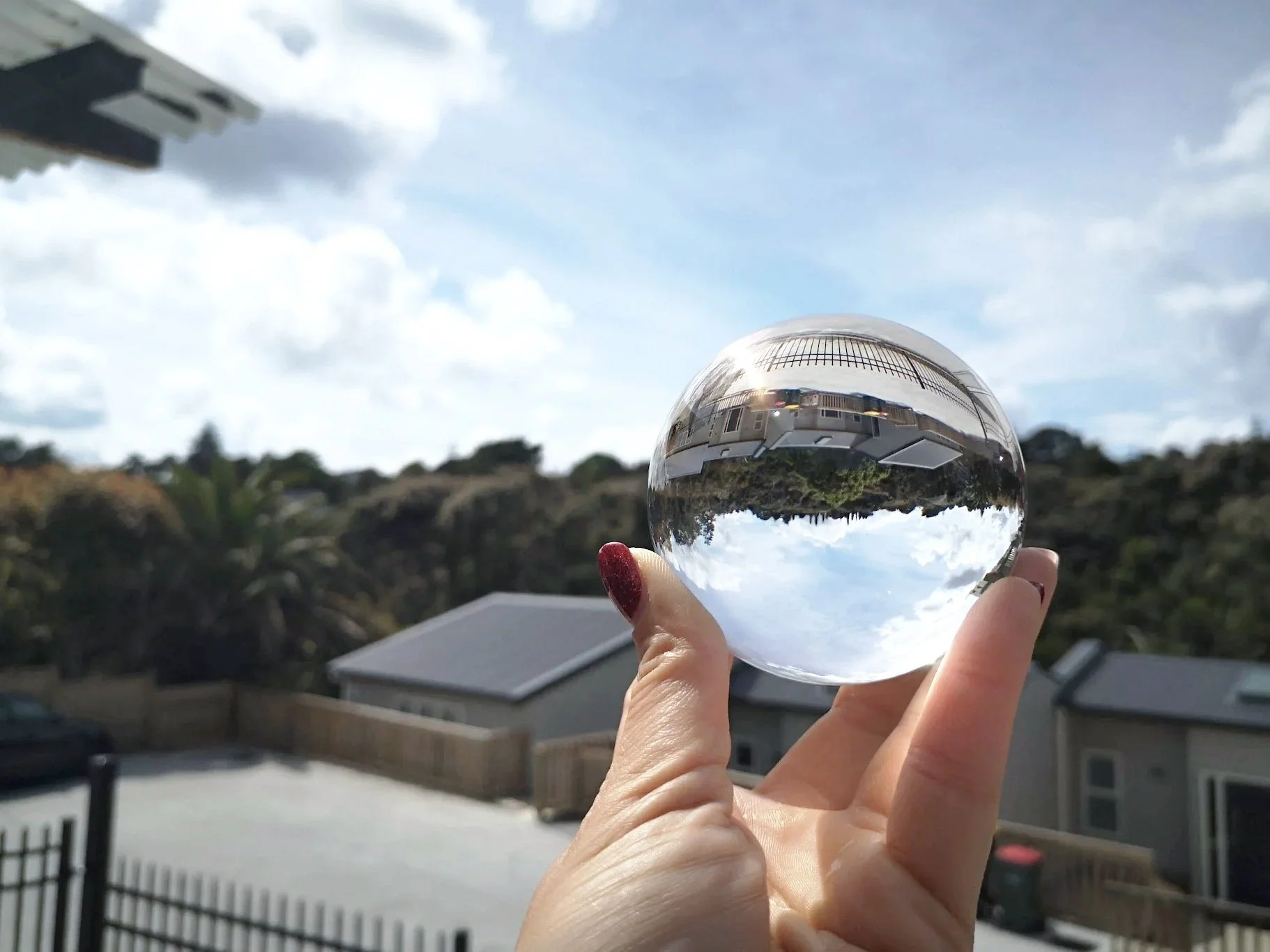 A hand with red-painted nails holds a clear glass sphere outdoors, reflecting a blue sky with clouds and buildings in the background.