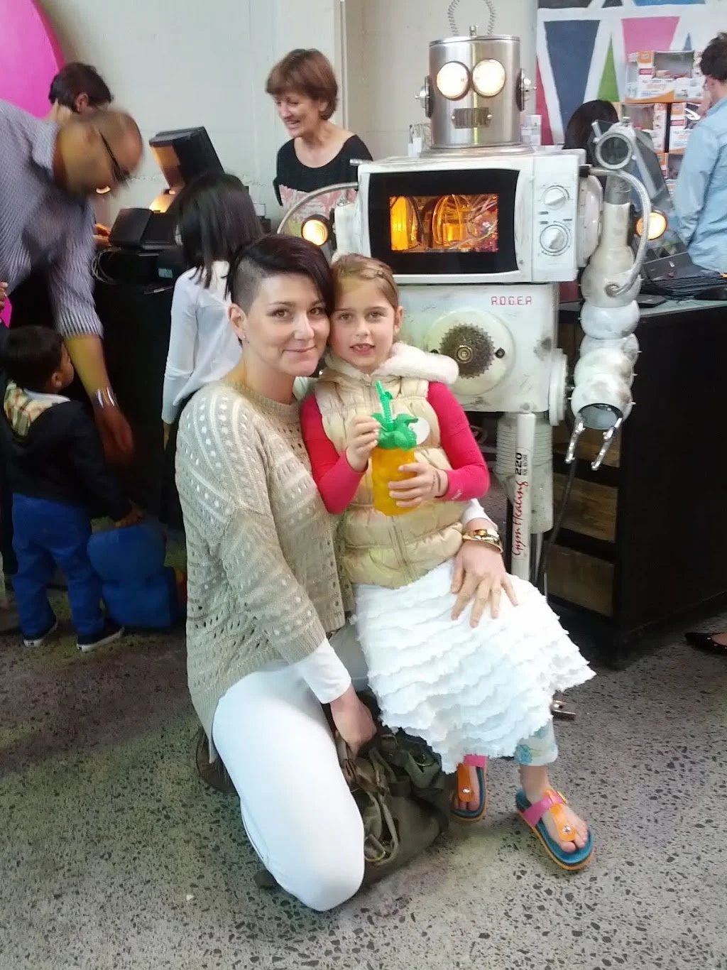 A woman and a girl smiling and posing in front of a robot in a busy indoor setting with several people, some children, in the background.