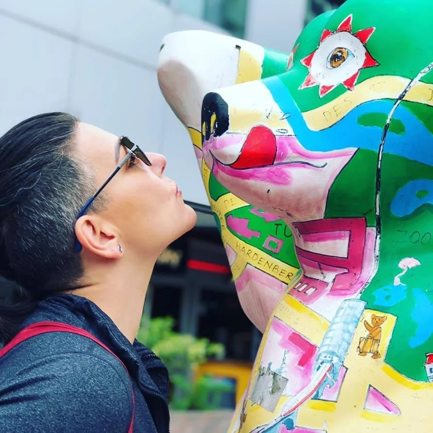 Person with short dark hair and glasses leaning toward a colorful bear-shaped sculpture, appearing to kiss its nose.