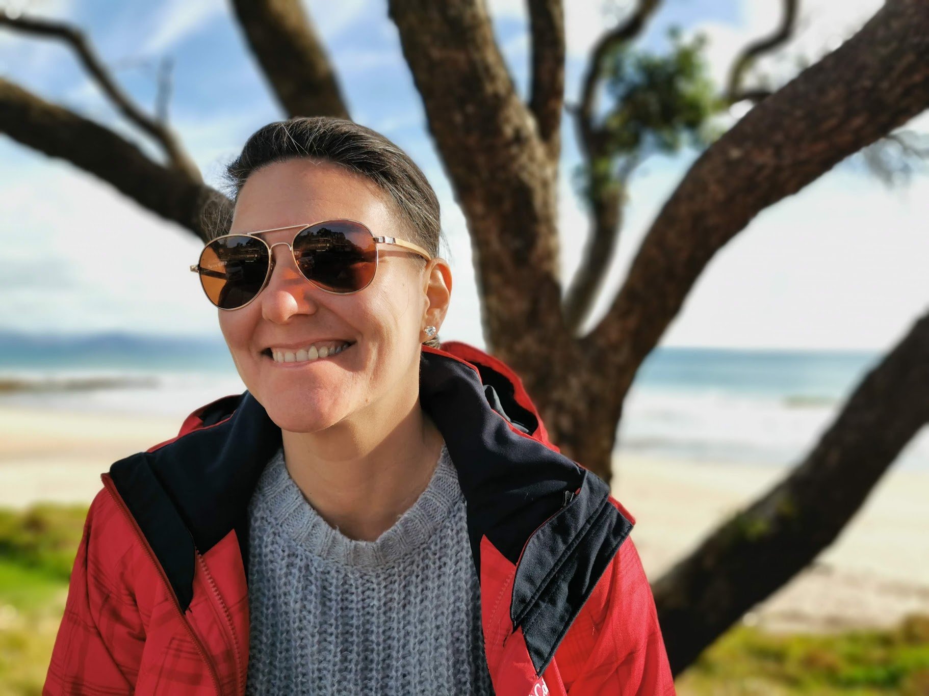 Kati Ohens smiling outdoors with sunglasses, wearing a gray sweater and a red and black jacket, standing in front of a tree near a beach with ocean in the background.