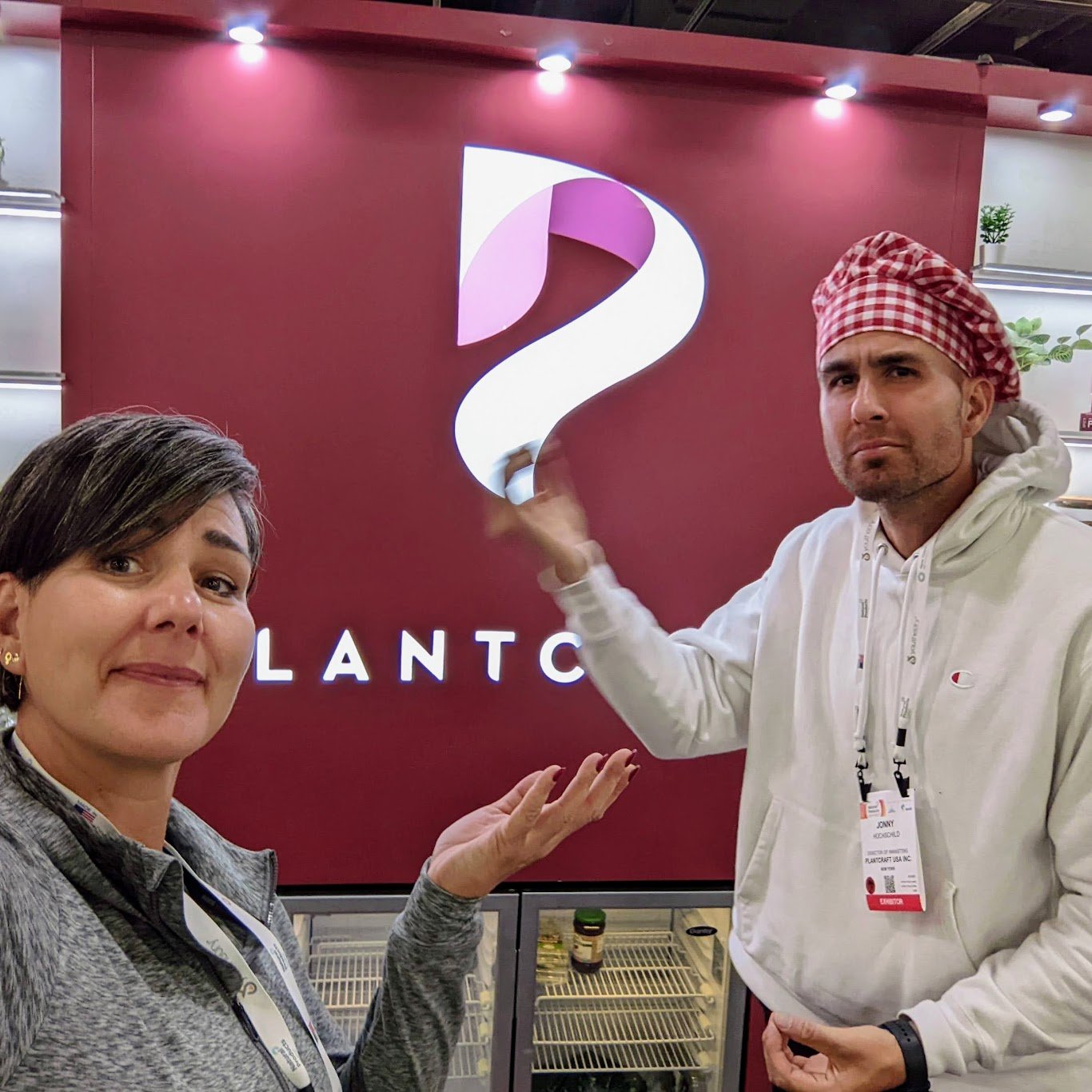Two people standing in front of a red wall with a large illuminated question mark logo and the word 'PLANTC' visible below. The woman on the left has short dark hair and is wearing a gray jacket, smiling and pointing at the logo. The man on the right