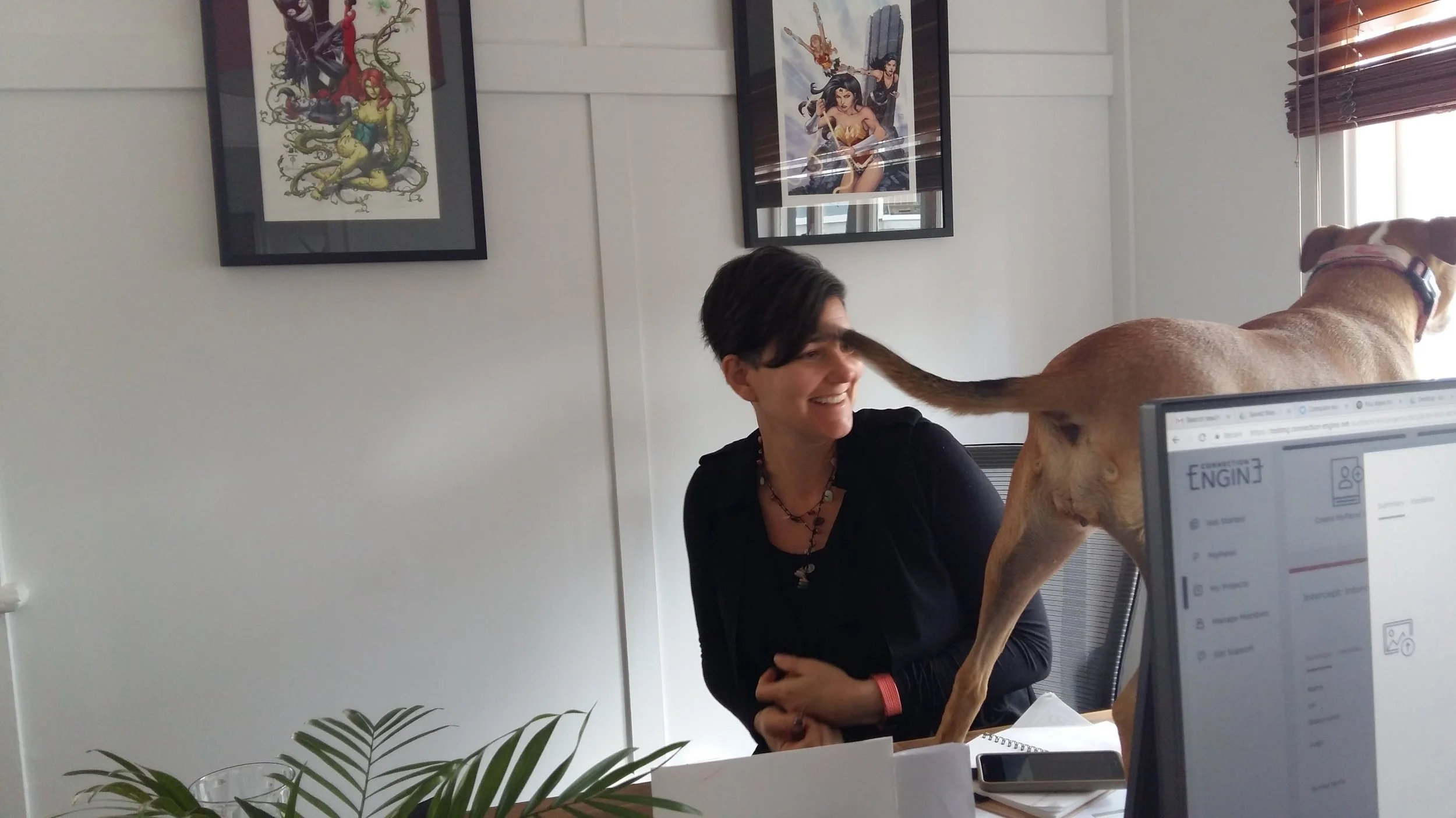 Kati Ohens smiling at her dog Roxy, who is sniffing a computer monitor on her desk in an office setting with framed artwork on white walls and a window with blinds.