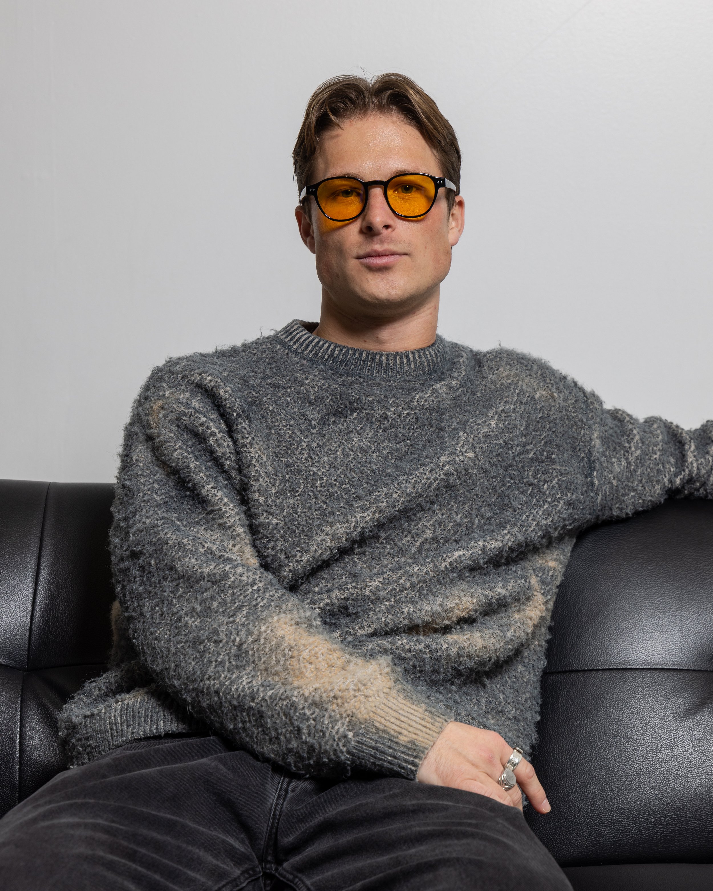 A young man with light skin and brown hair sitting on a black leather sofa, wearing yellow-tinted glasses, a gray textured sweater, and black pants.