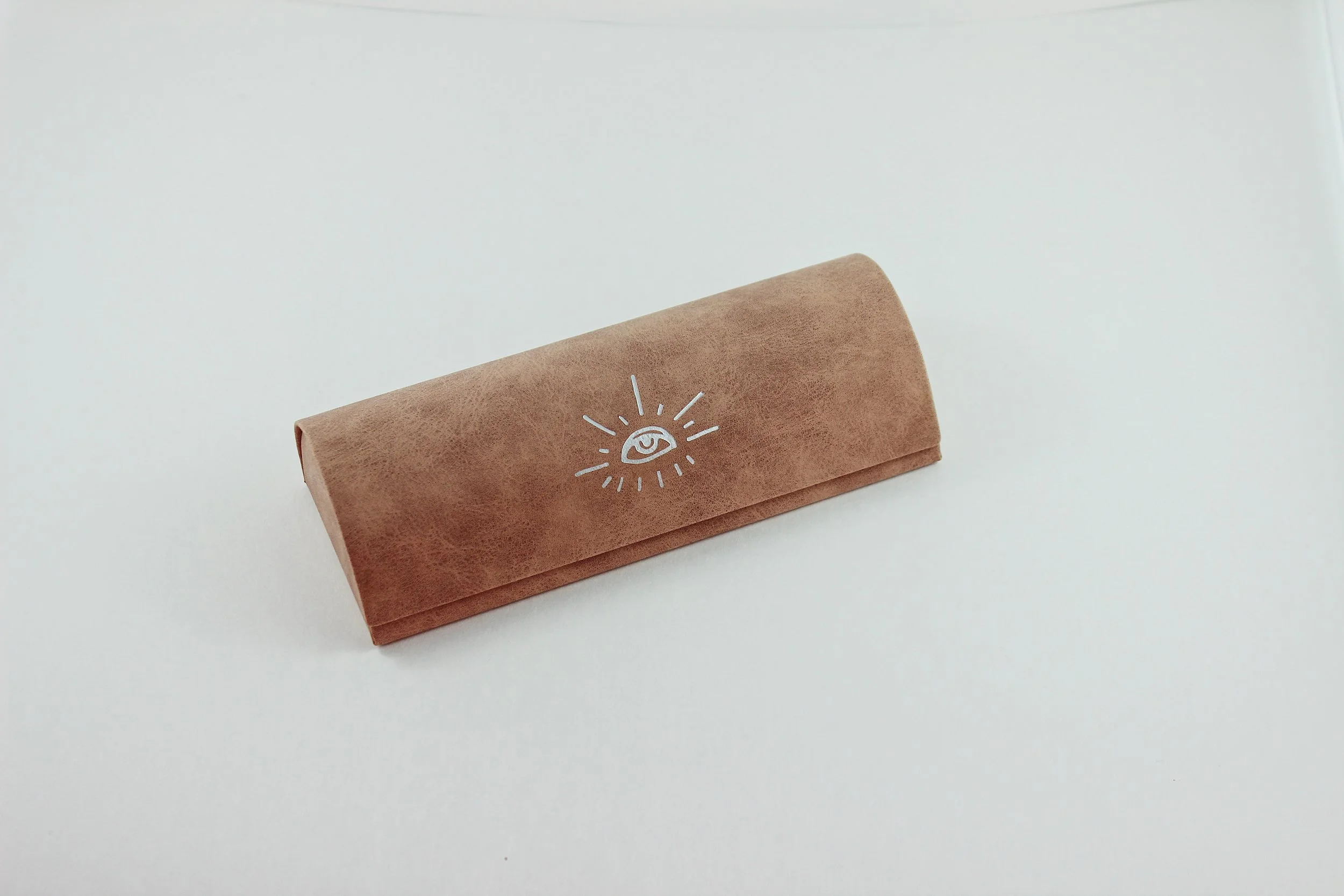 Brown handcrafted eyeglasses case with a white eye and rays design on the lid, placed on a white surface.