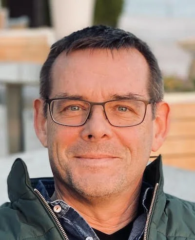 Close-up of a middle-aged man with dark hair, glasses, and a slight smile, wearing a dark green jacket outdoors.