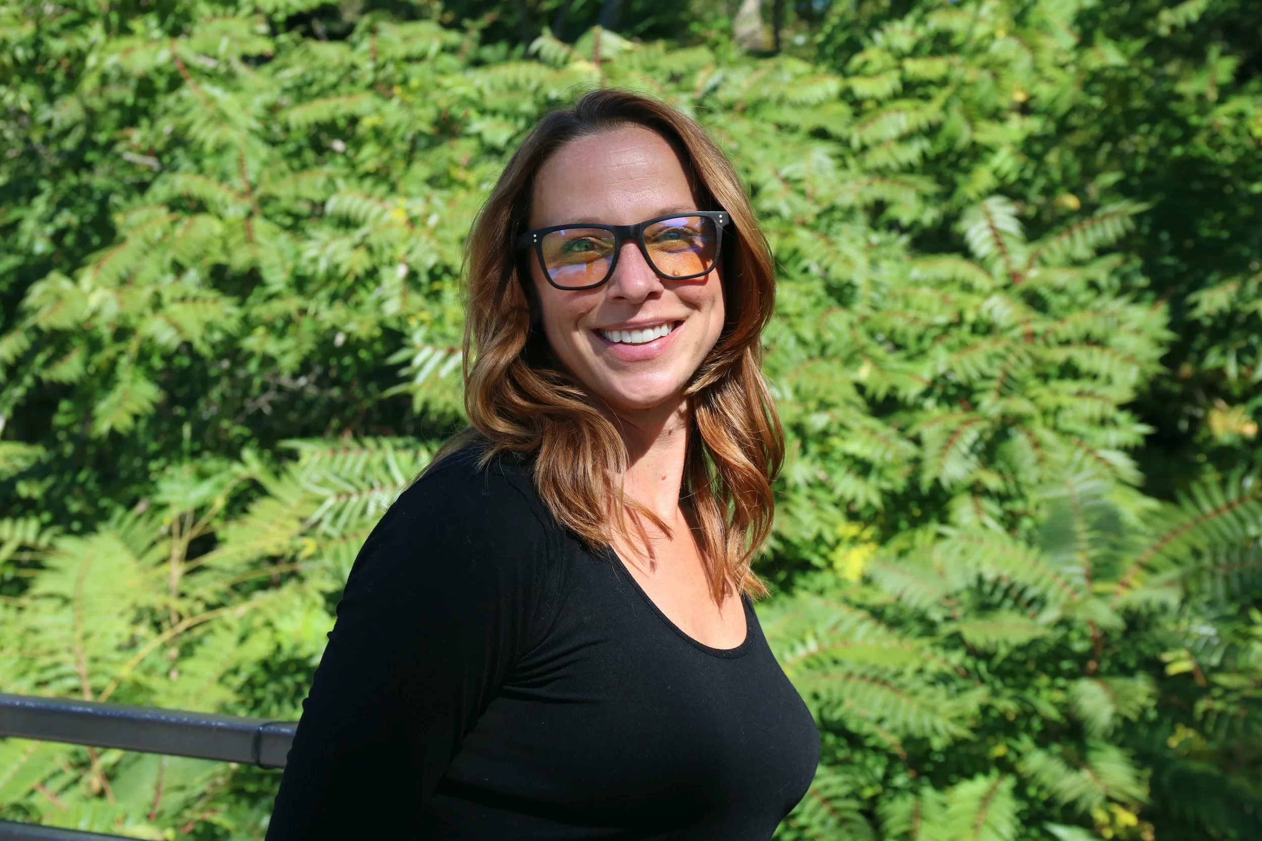 A woman with shoulder-length brown hair, wearing a black top and sunglasses, smiling outdoors in front of green foliage.