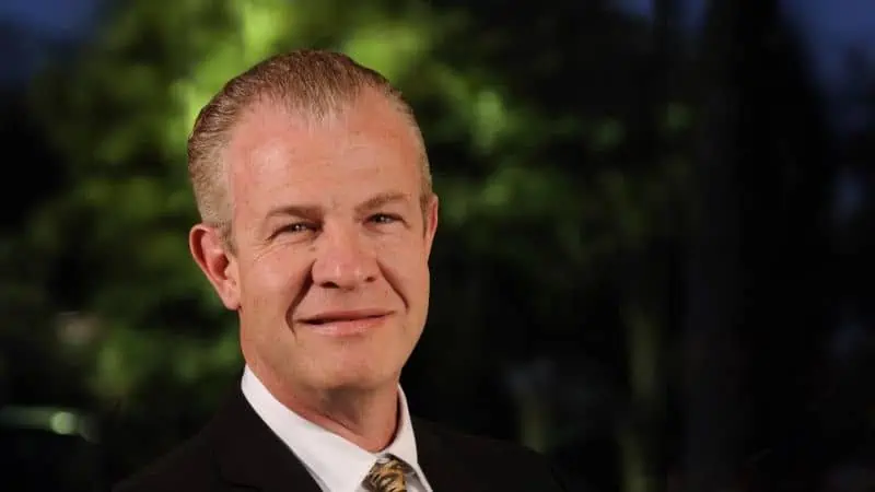 A man with short, slicked-back hair wearing a dark suit, white shirt, and a patterned tie, smiling slightly in front of a blurred outdoor background.