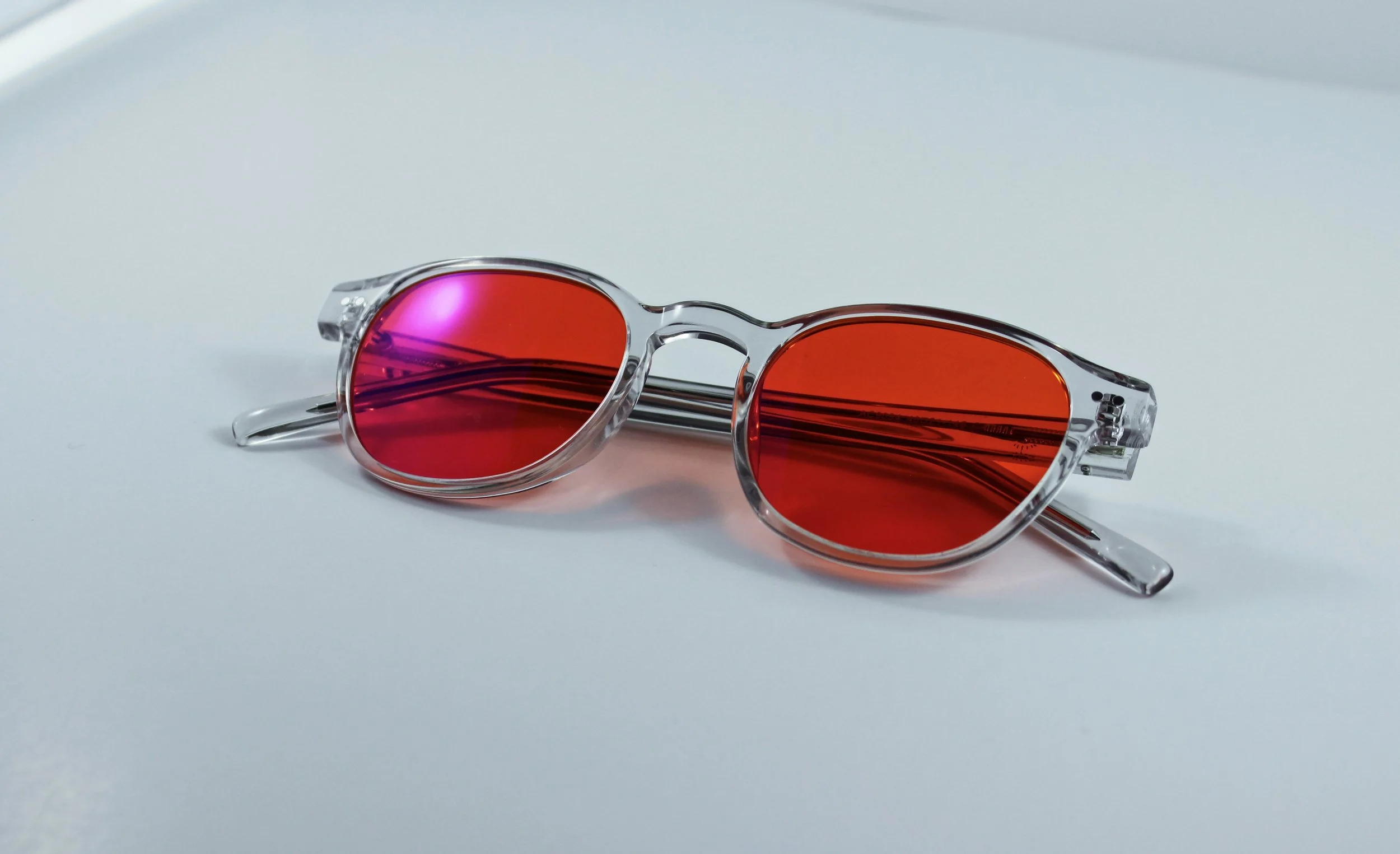 Pair of transparent blue light glasses with red mirrored lenses on a white surface.