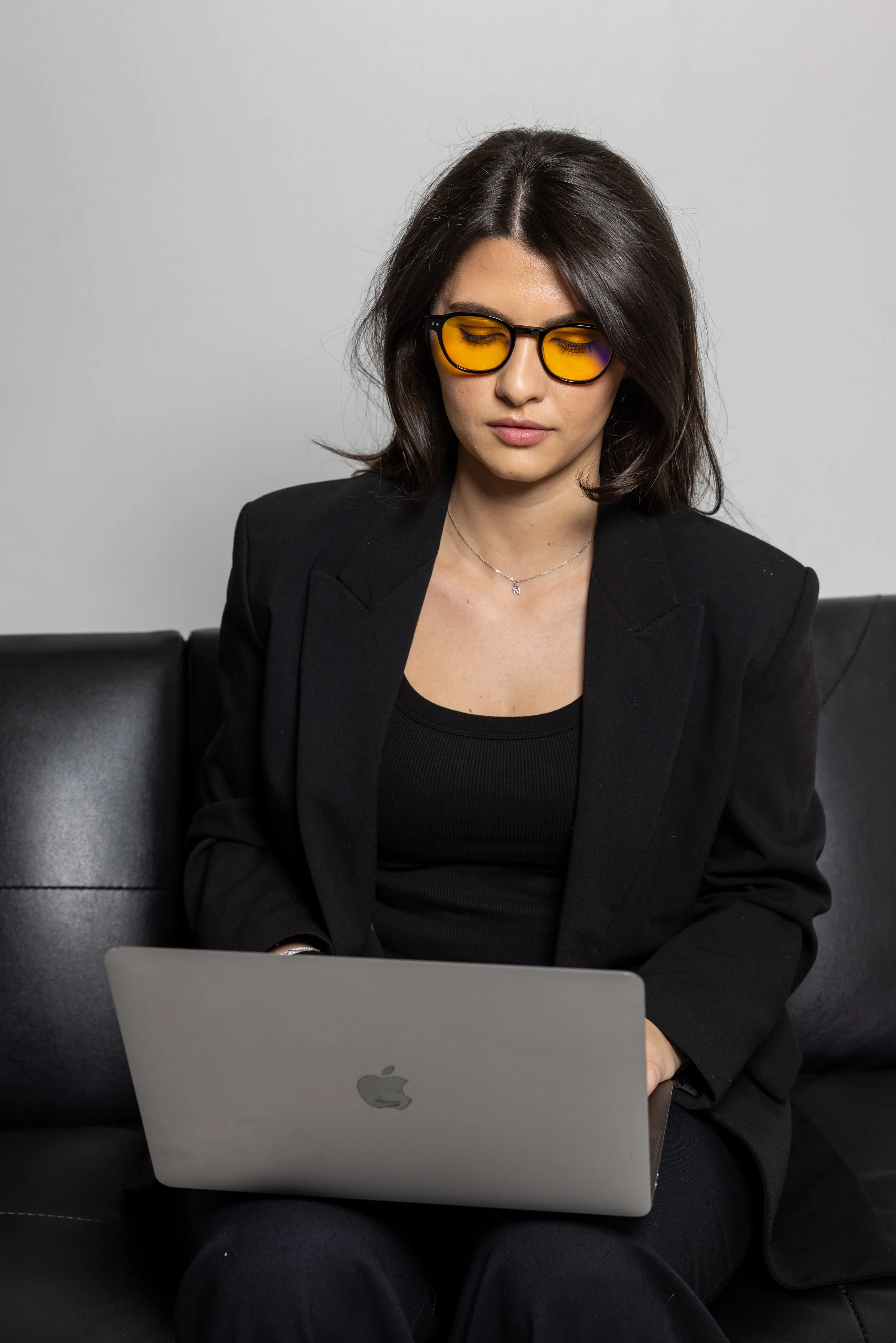 A woman with dark brown hair and glasses with yellow lenses working on a silver Apple MacBook laptop while sitting on a black leather couch.