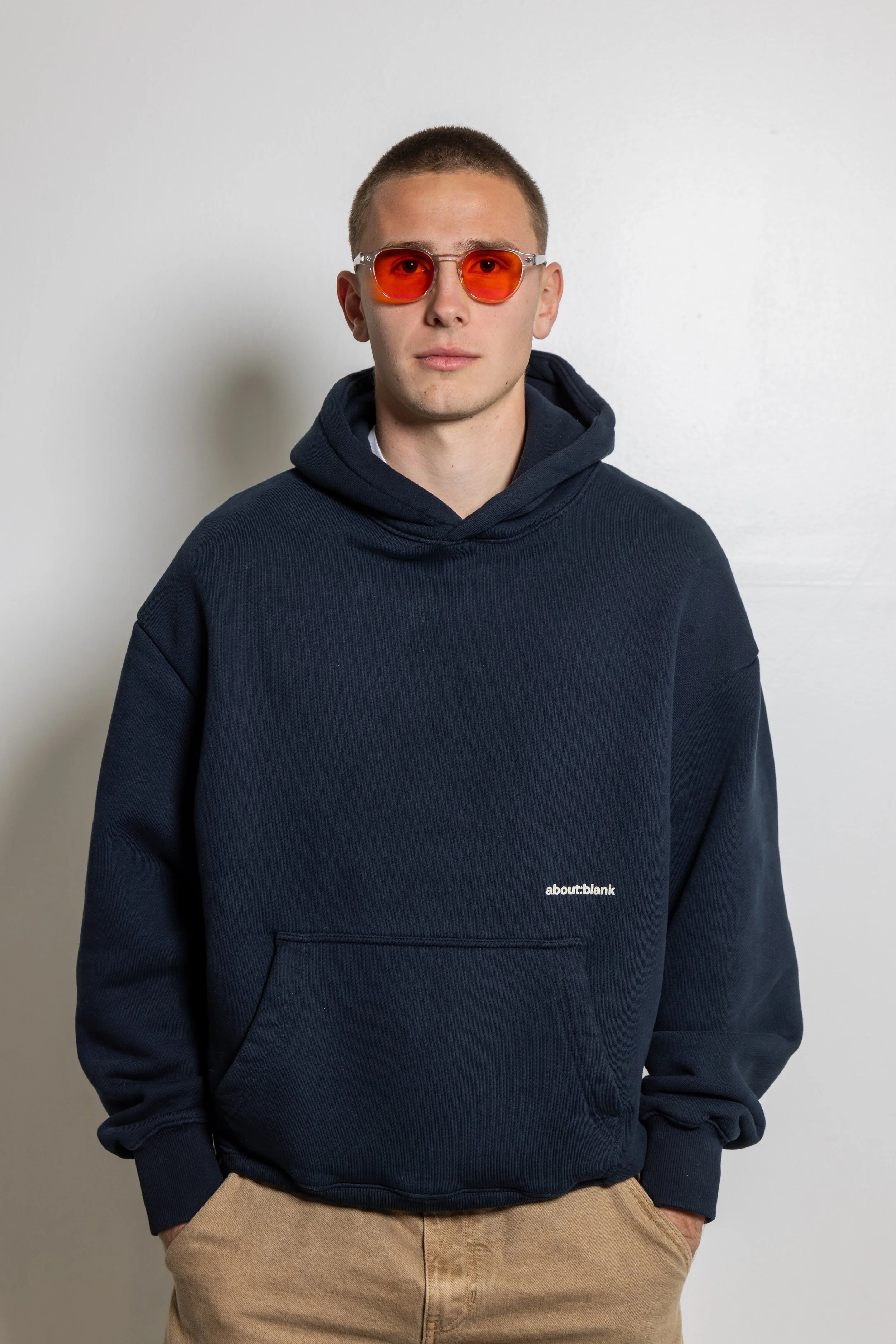 A young man with short hair wearing orange blue light glasses, a navy blue hoodie with small white text that reads 'about:blank,' and beige pants standing against a white wall.