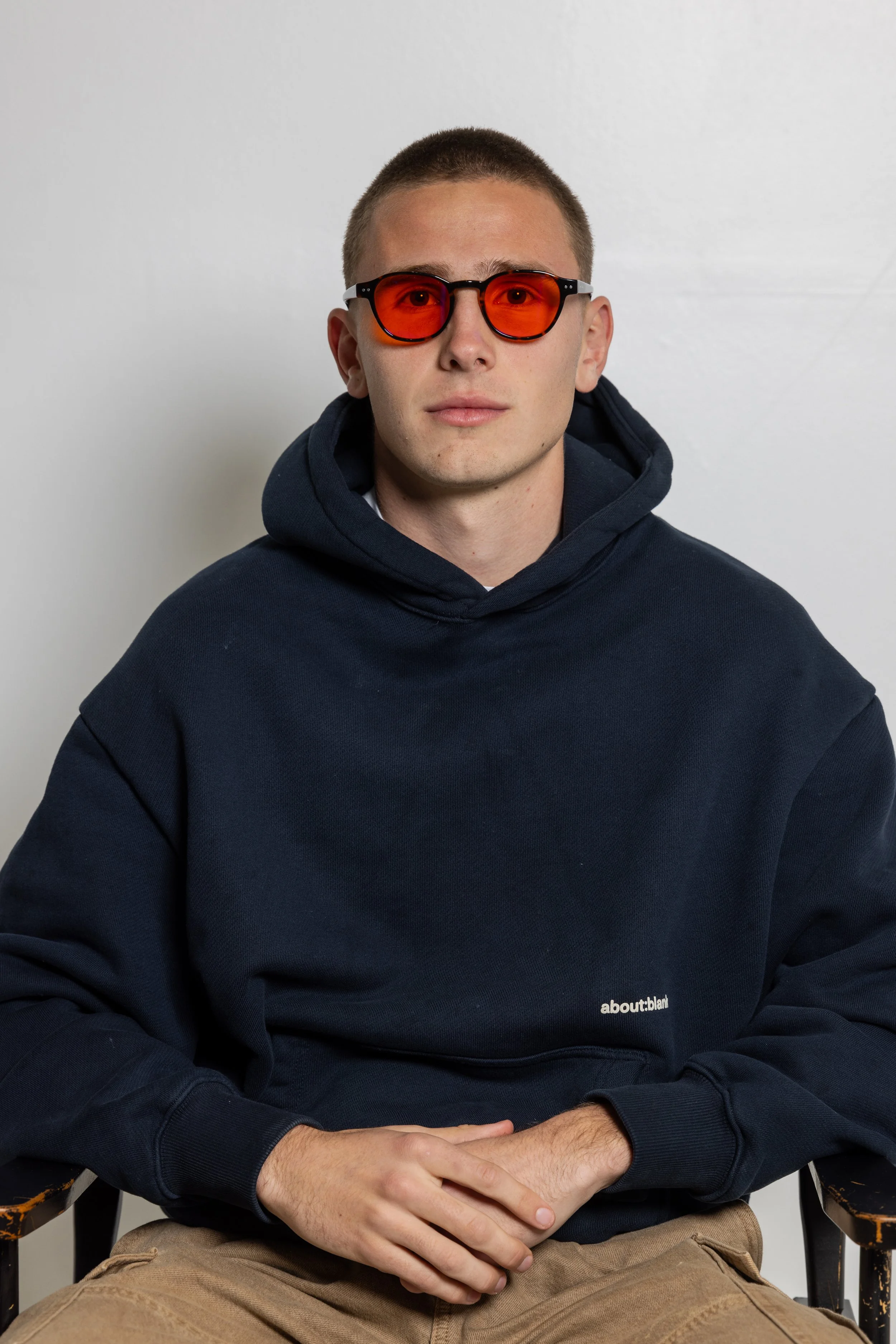 A young man with short hair wearing red-tinted blue light glasses, a navy hoodie with the words 'about:blank' on it, and tan pants, sitting against a plain white wall.