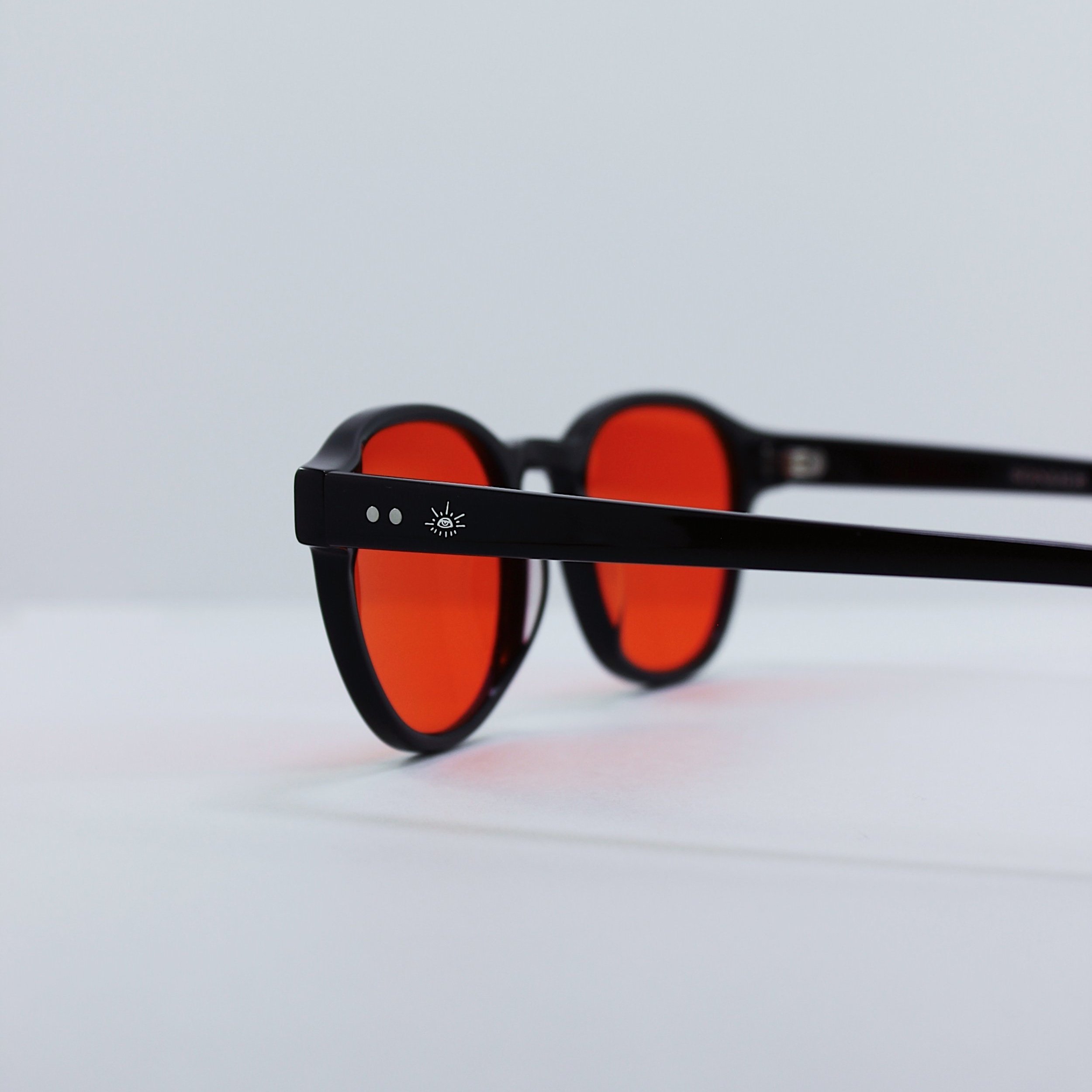 Black blue light glasses with red lenses placed on a white surface, with a small icon of a sun and two dots on the temple arm.
