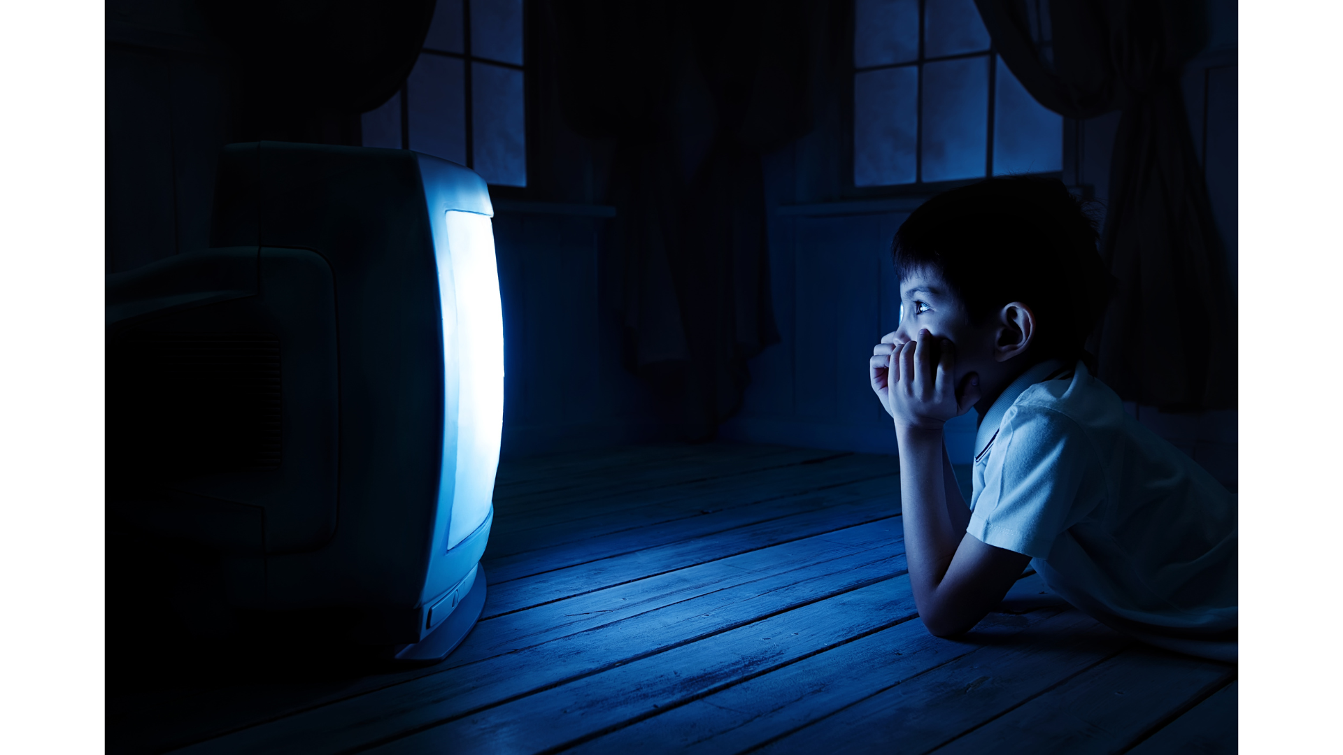 A young boy lies on the wooden floor in a dark room, watching television with a bright screen illuminating his face.
