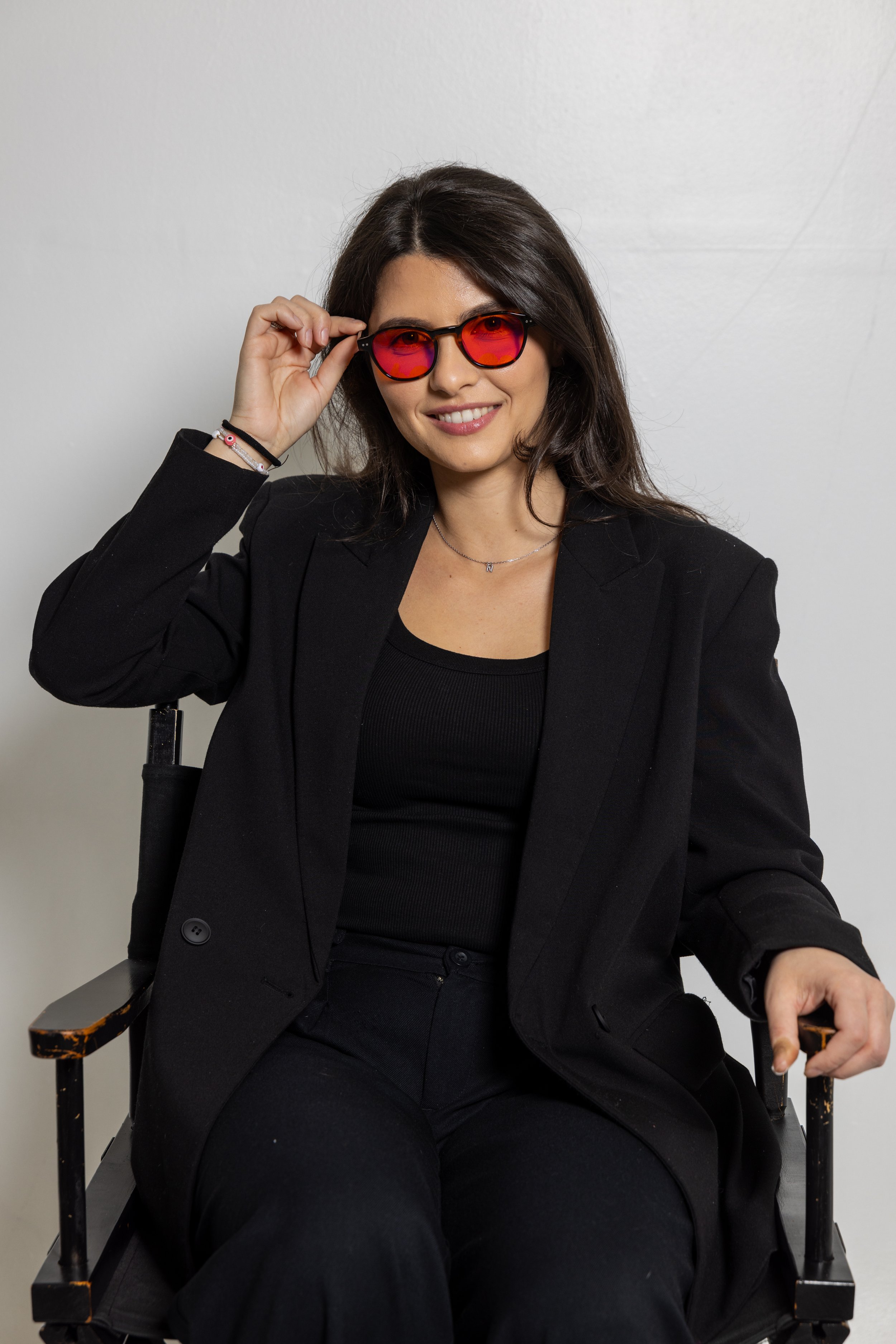 A young woman with long dark hair wearing a black blazer, black top, and sunglasses with red lenses, sitting in a black chair against a plain white wall, smiling at the camera.