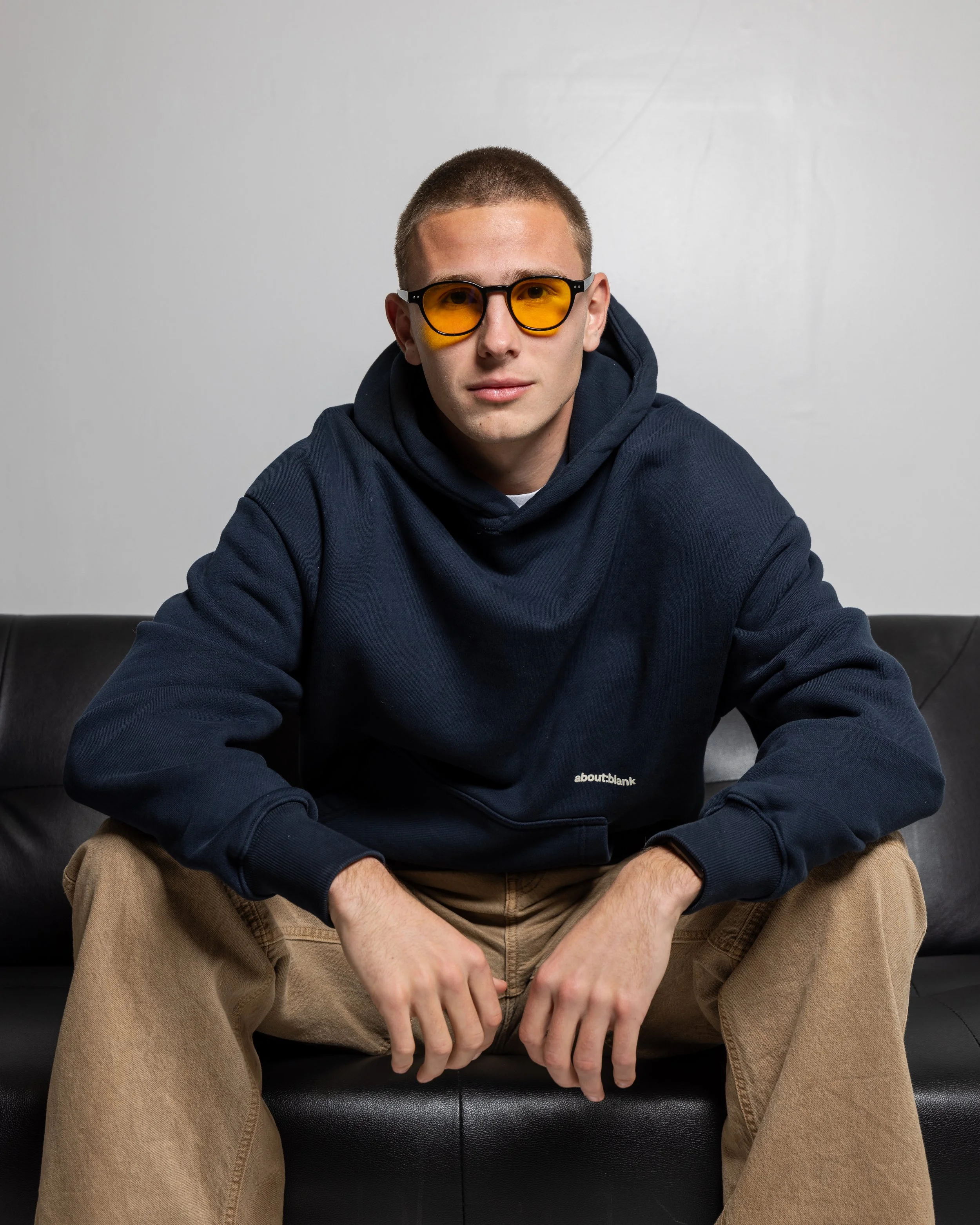 Young man sitting on a black couch wearing a navy hoodie, beige pants, and yellow-tinted sunglasses, with a neutral expression against a plain gray background.