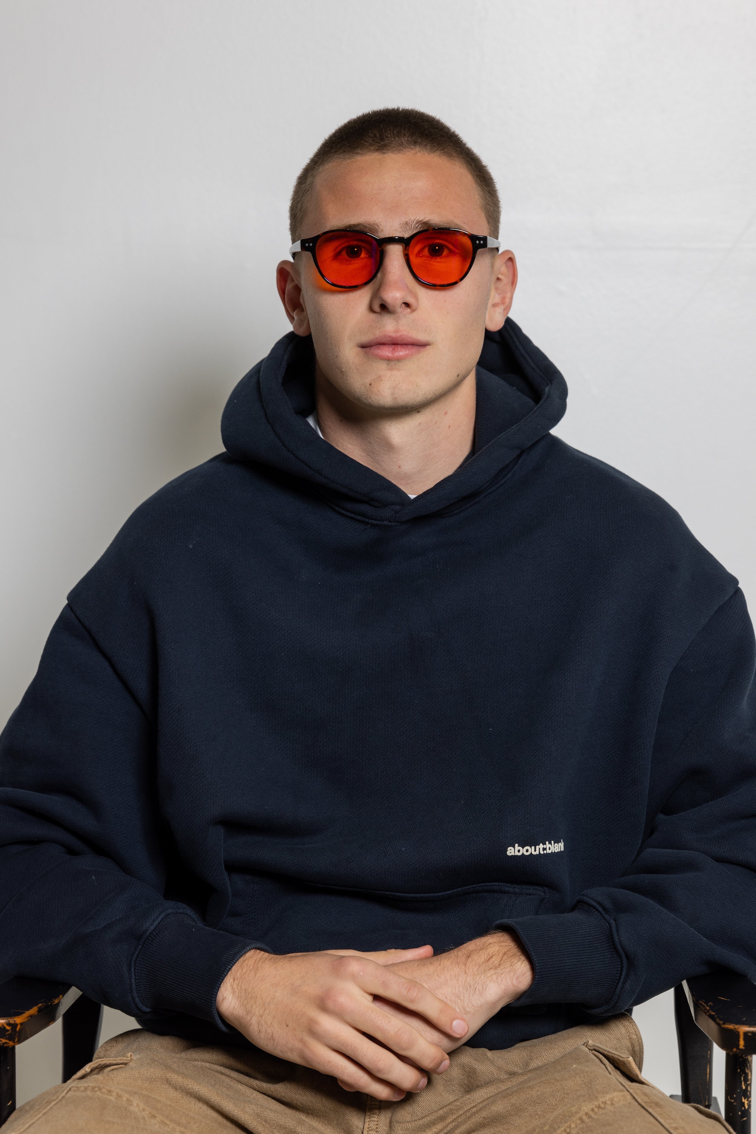 A young man wearing red-tinted blue light glasses glasses, a navy blue hoodie with the text 'about:blank,' and beige pants, sitting with hands clasped in front of him against a plain white wall.