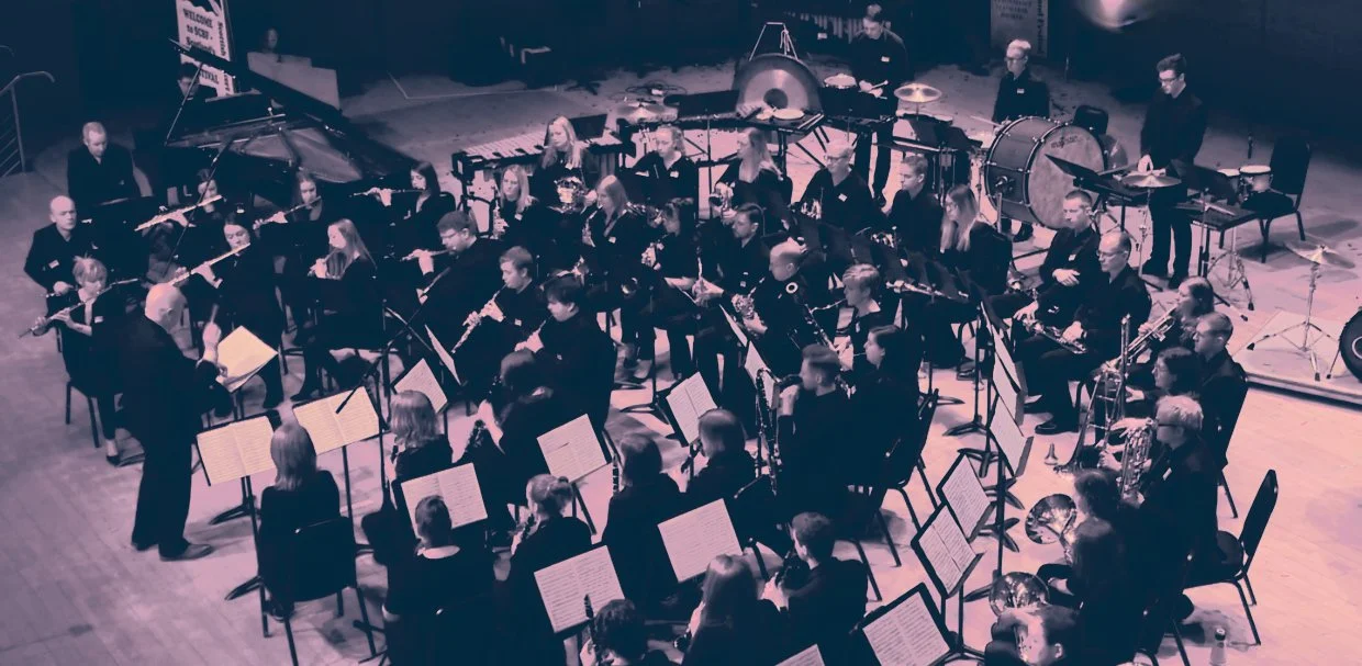 An orchestra performing on stage with musicians playing various instruments including strings, woodwinds, brass, percussion, and a grand piano, conducted by a person standing in front.