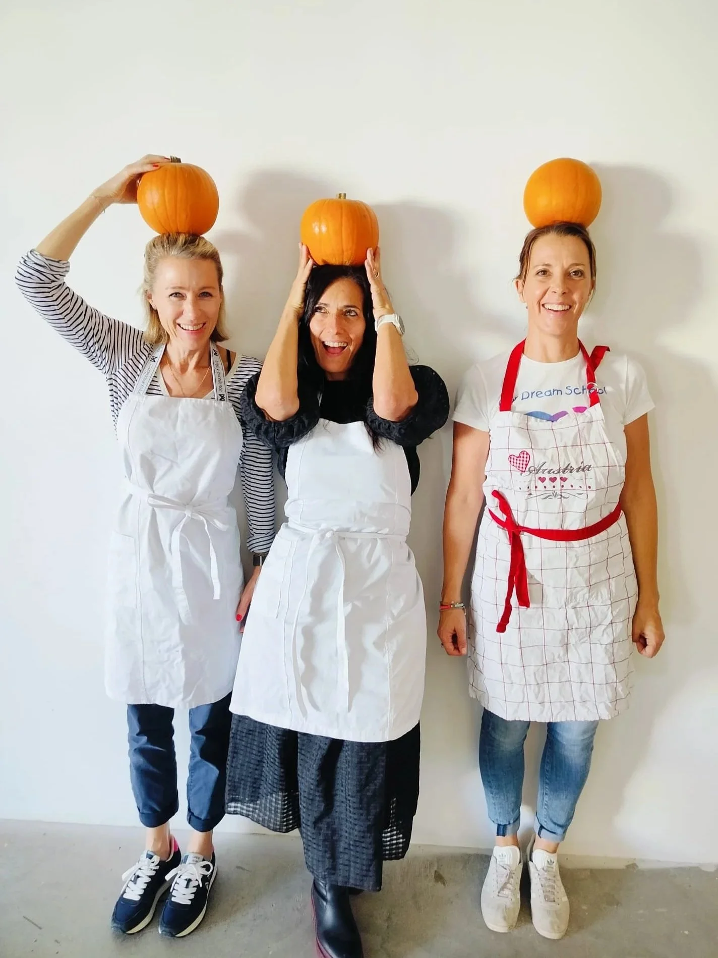 Three women standing against a plain wall, each balancing a pumpkin on their head. They are smiling and wearing aprons, with the middle woman wearing a black top and the other two wearing white tops. All are dressed casually and appear to be having fun.