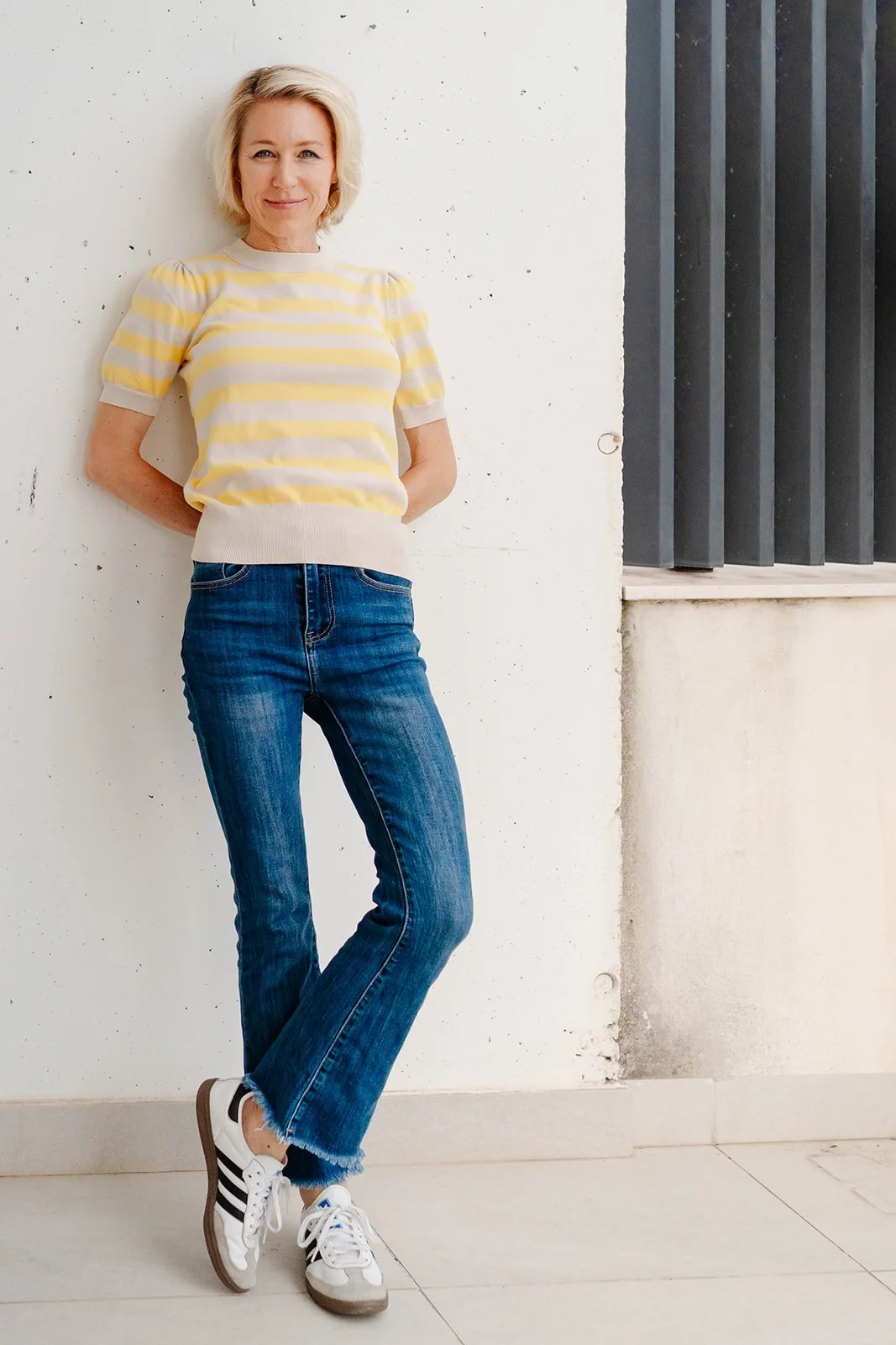 Woman standing against a white wall with a black metal grate nearby, wearing a yellow and white striped sweater, blue jeans, and sneakers.