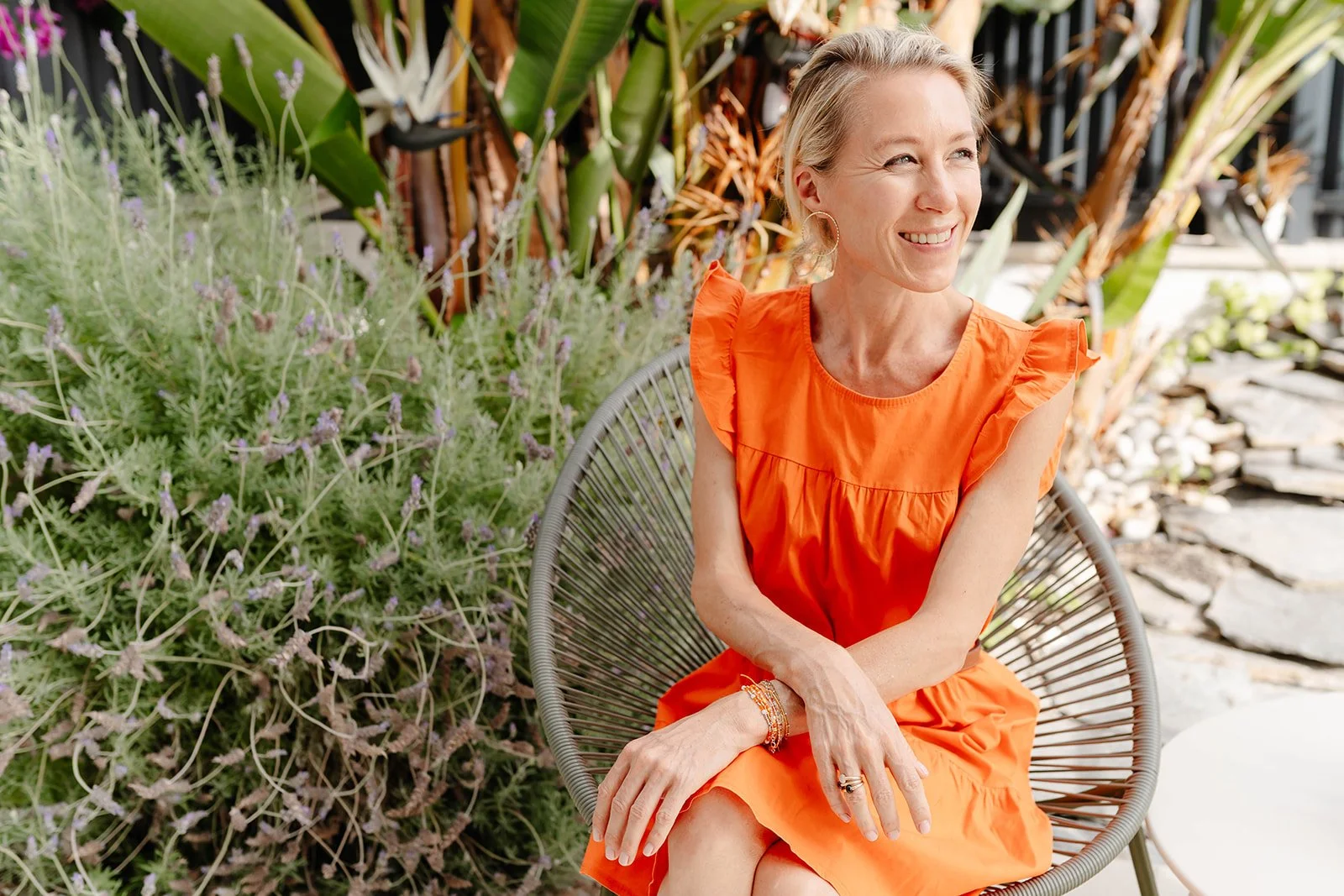 A woman in an orange dress sitting on a round chair outdoors, smiling and looking to her right, surrounded by green plants and garden decor.
