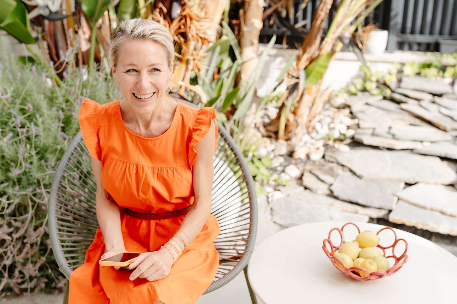 Smiling woman in an orange dress sitting outside on a modern chair with a tablet in her hand, next to a white table with a fruit bowl of yellow lemons, with plants and stone pathway in the background.