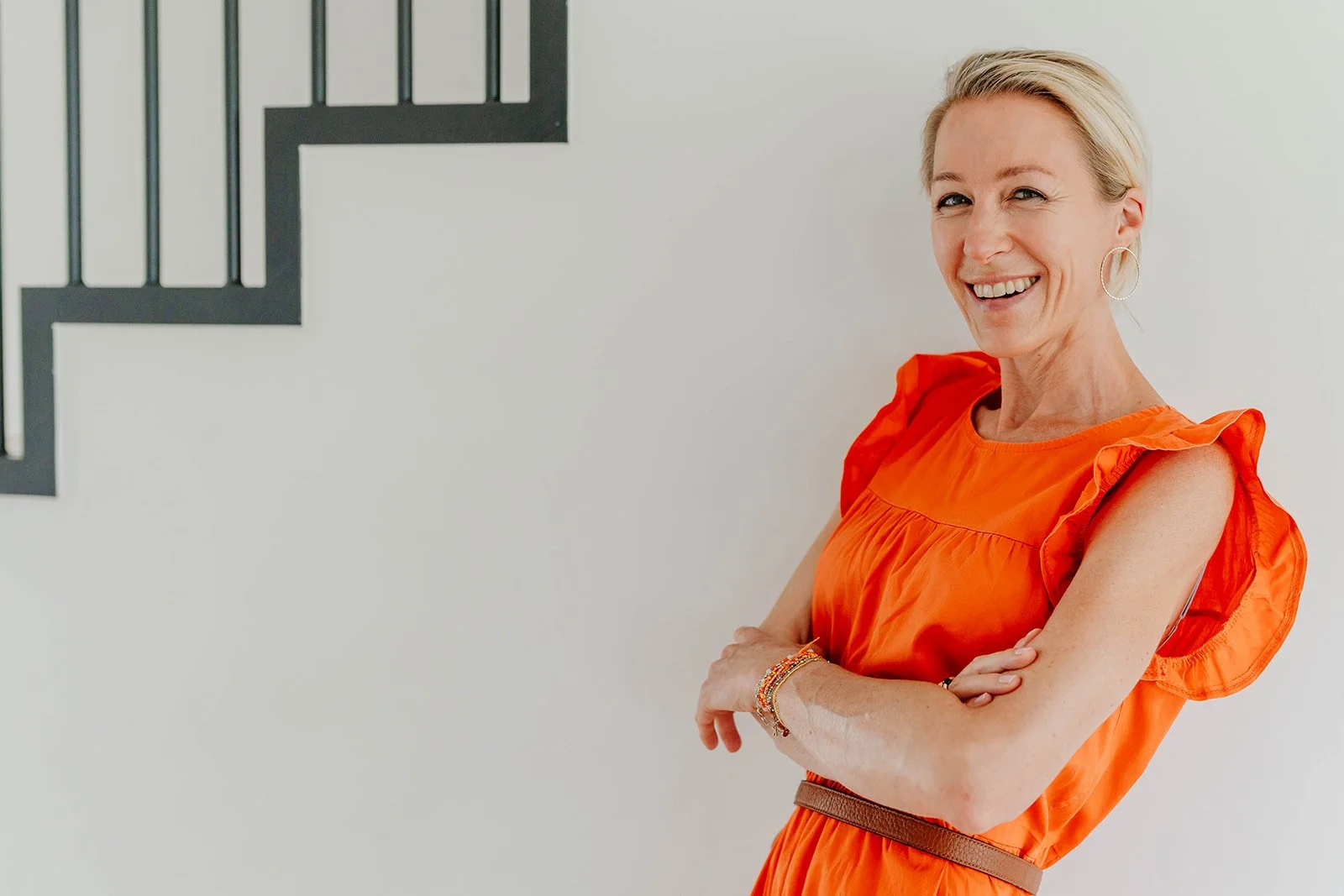 Smiling woman in an orange ruffled dress standing with arms crossed in front of a staircase