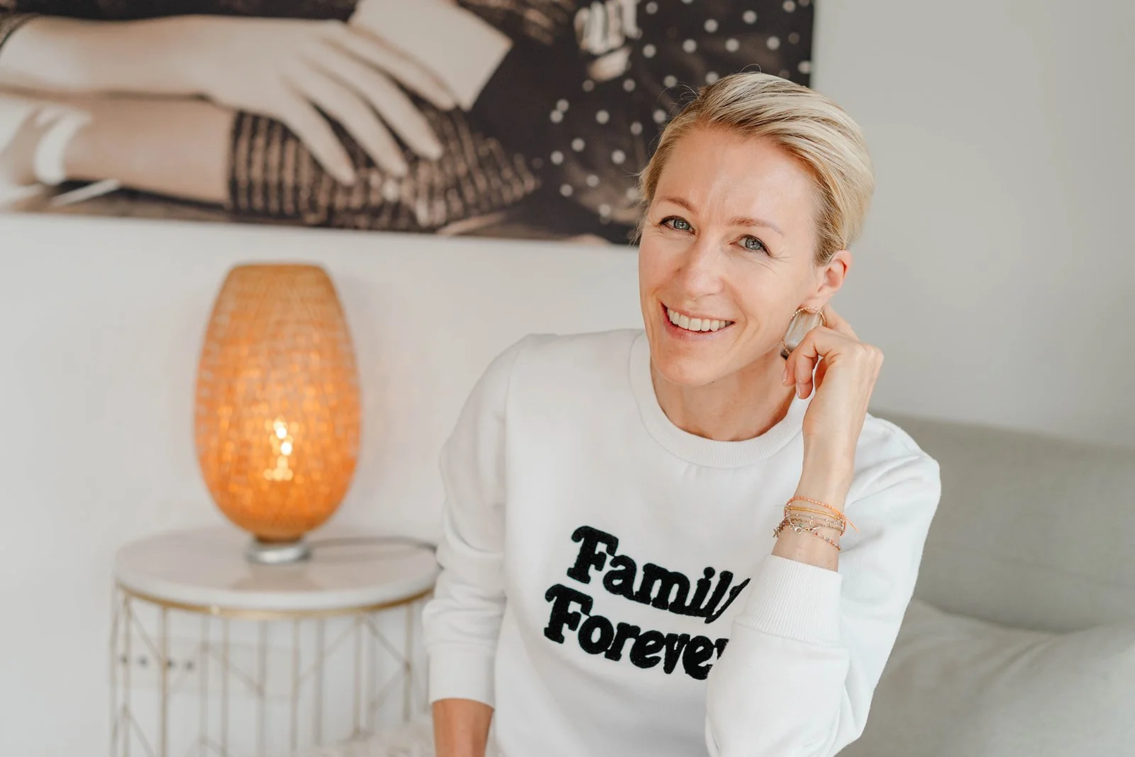 A woman with short blonde hair smiling, wearing a white sweatshirt with black text that reads "Family Forever," sitting in a living room with a side table, a woven lamp, and a black and white framed photo on the wall.