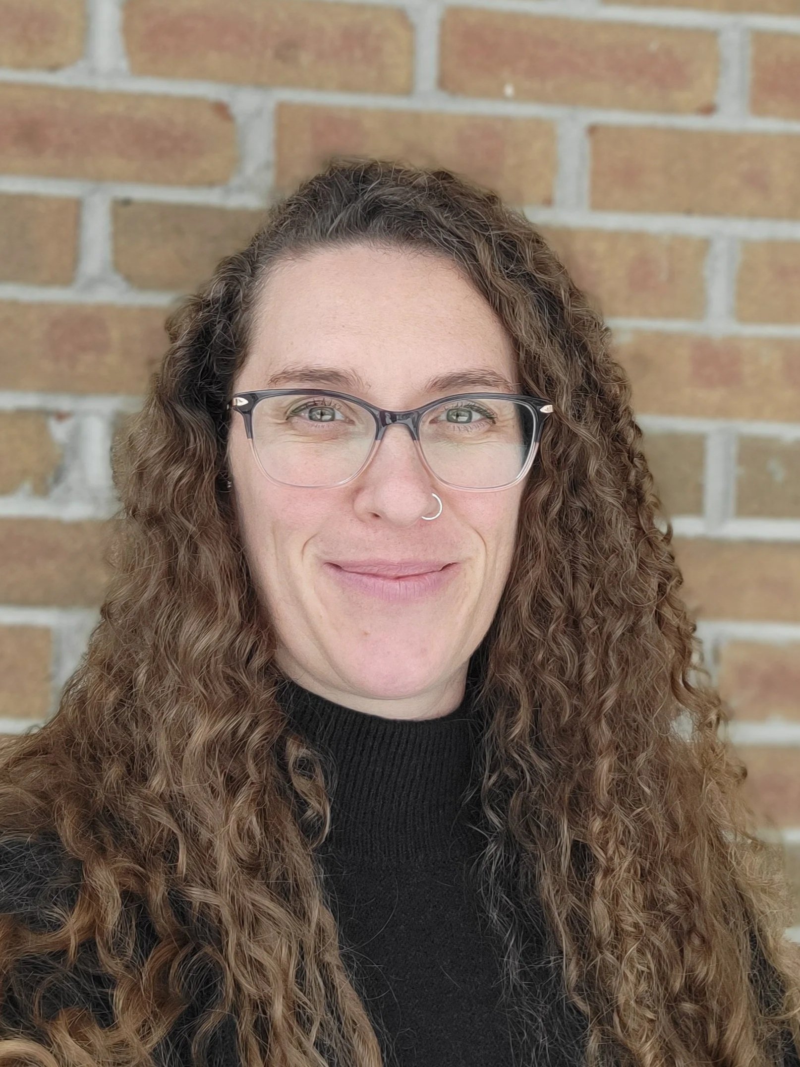 A woman with curly brown hair, wearing glasses and a nose ring, smiling in front of a brick wall.