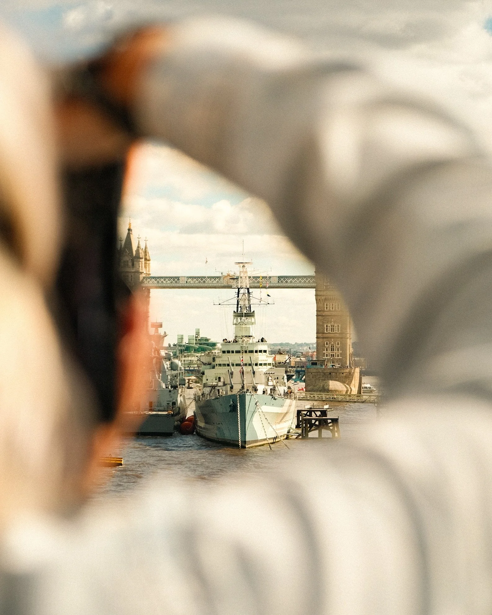 A view of a harbor with multiple boats docked, framed through a blurred foreground with buildings and a bridge in the background.