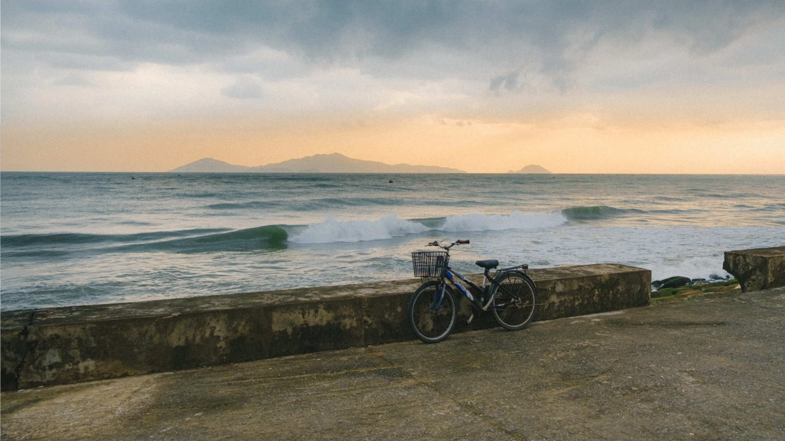 A bicycle with a basket parked on a concrete barrier near the beach, with ocean waves and distant islands under a cloudy sky.