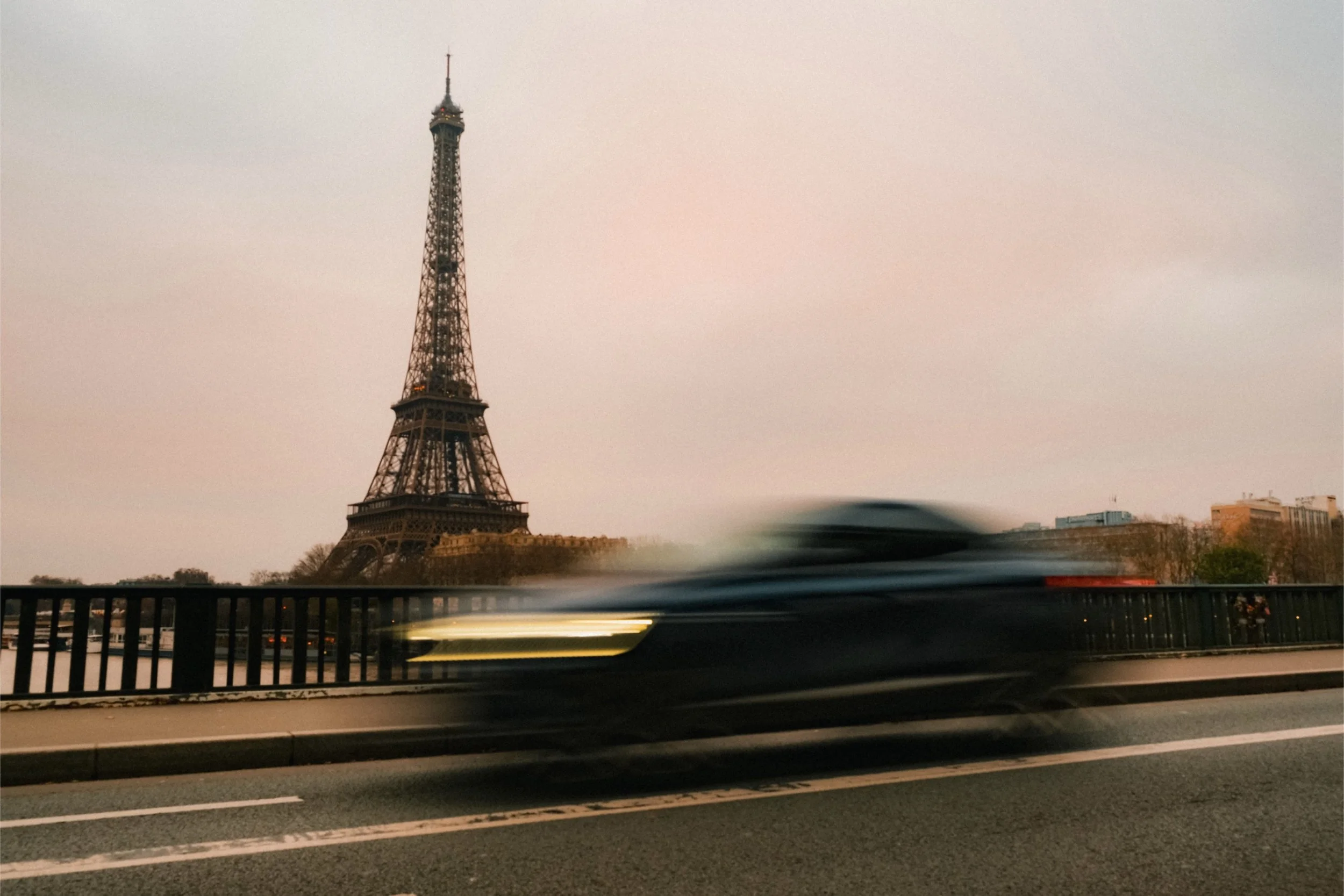The Eiffel Tower in Paris seen from a road with blurred moving vehicles in the foreground during dusk or early evening.