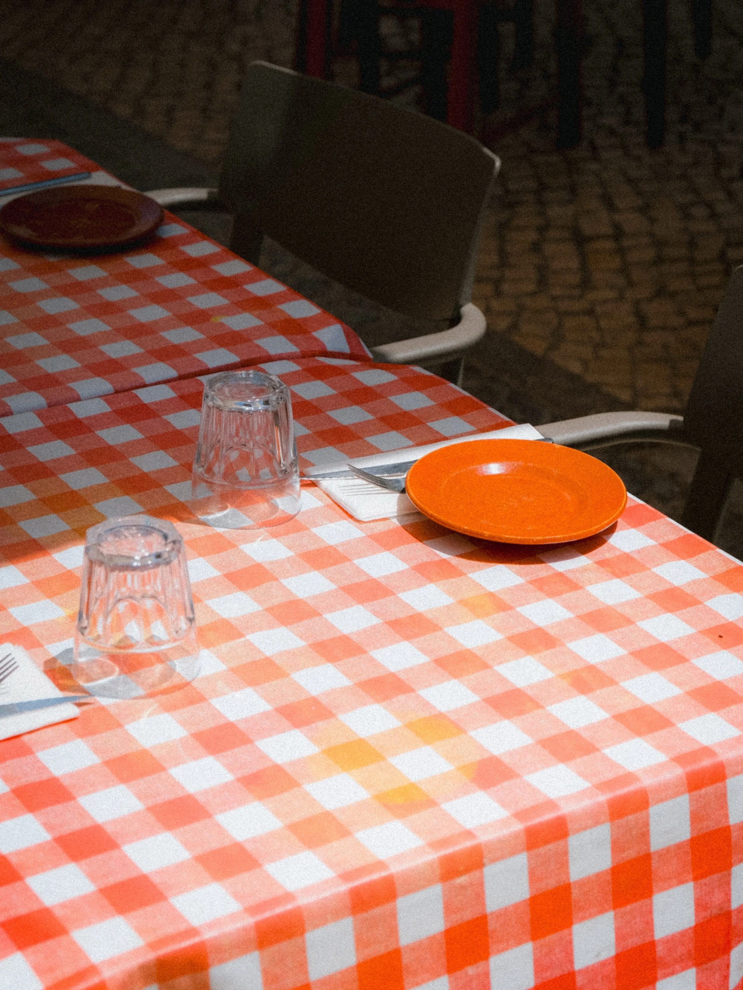 A table with a red and white checkered tablecloth, set with three upside-down clear glasses, two orange plates, and a utensil, with chairs in the background.