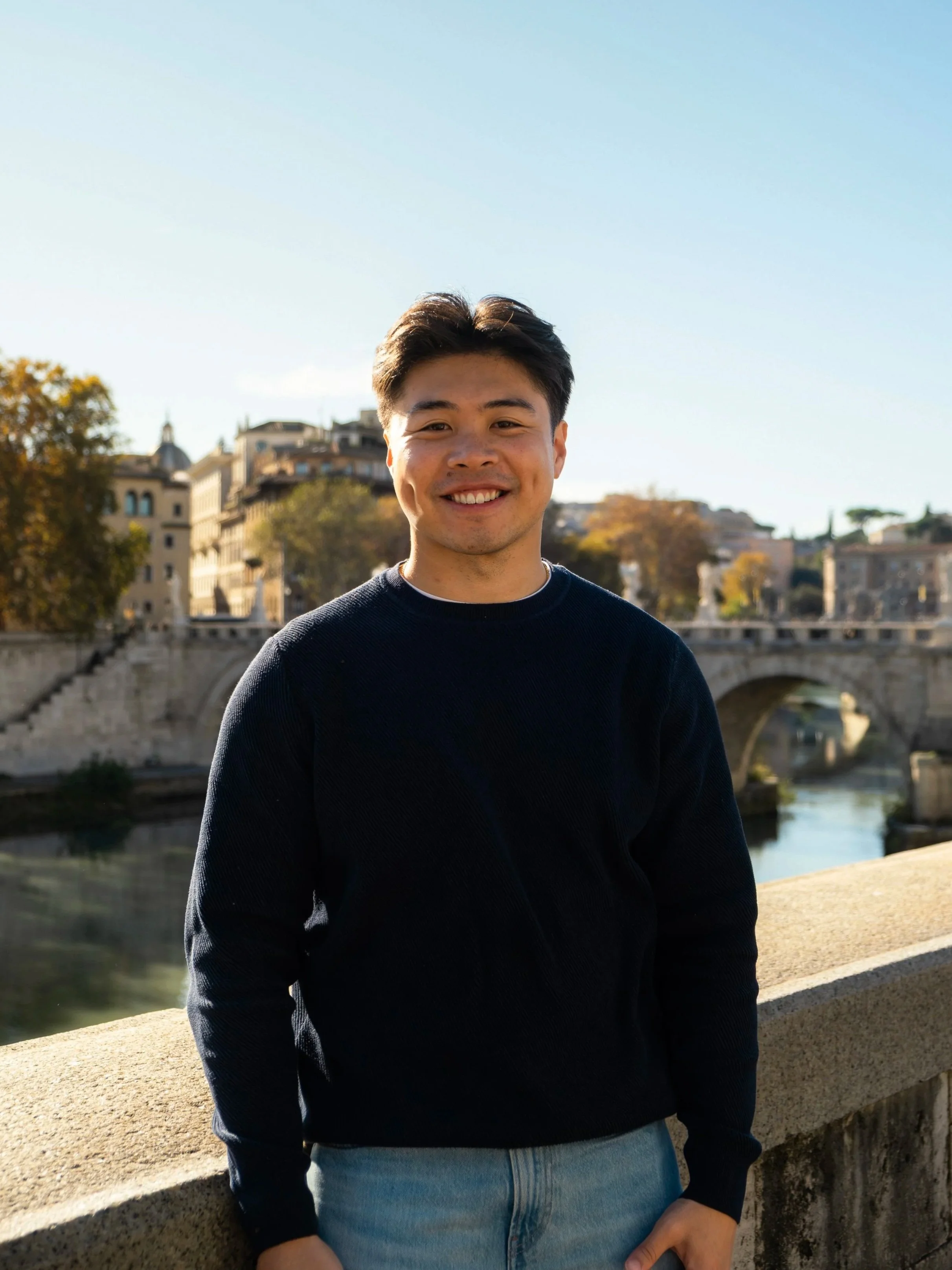 Young man smiling outdoors near a bridge with buildings and trees in the background. Professionally taken, it show cases Richard Thai, a photographer and content creator.
