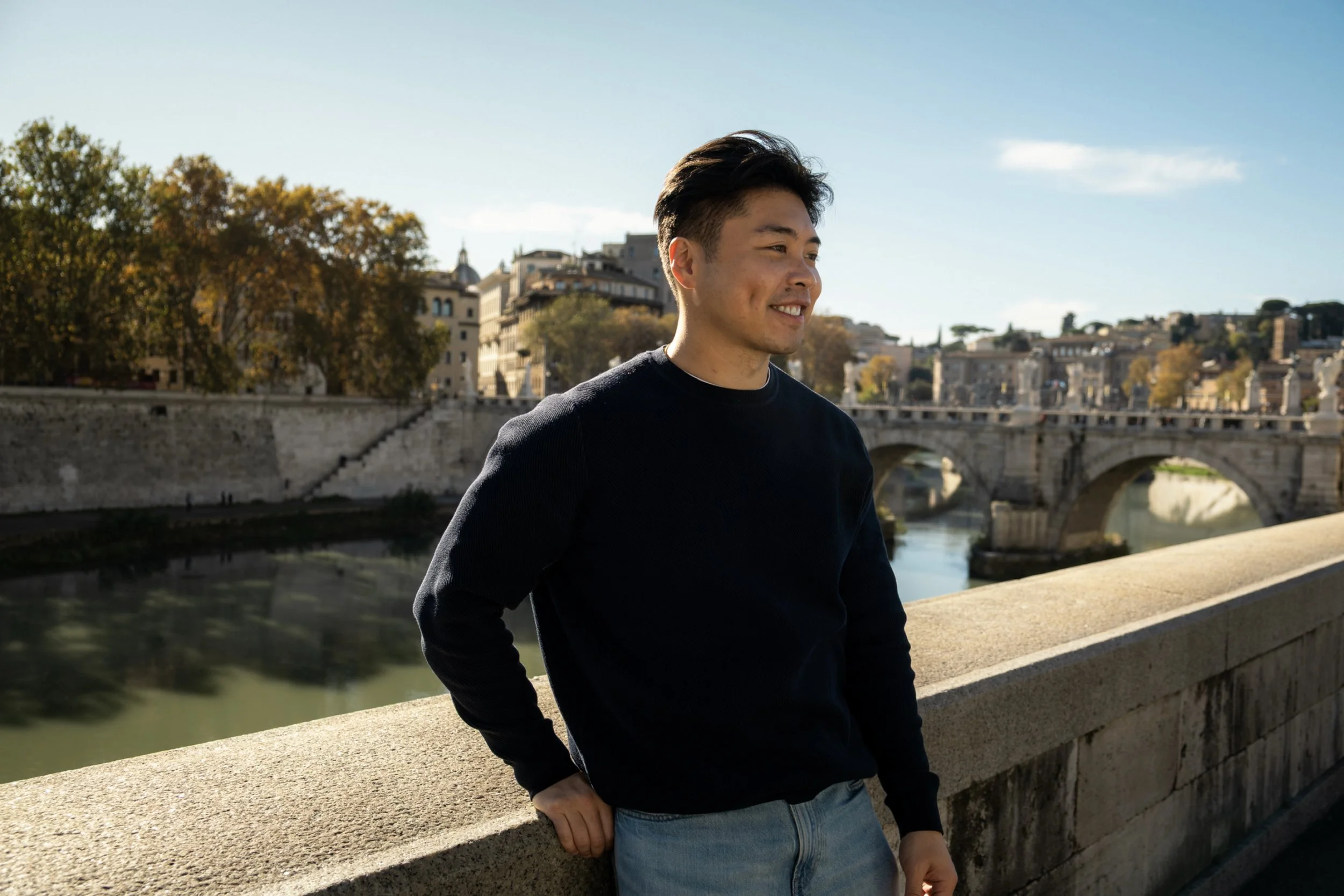 A young man with dark hair and a black shirt standing outdoors on a bridge, with city buildings and trees in the background on a sunny day.