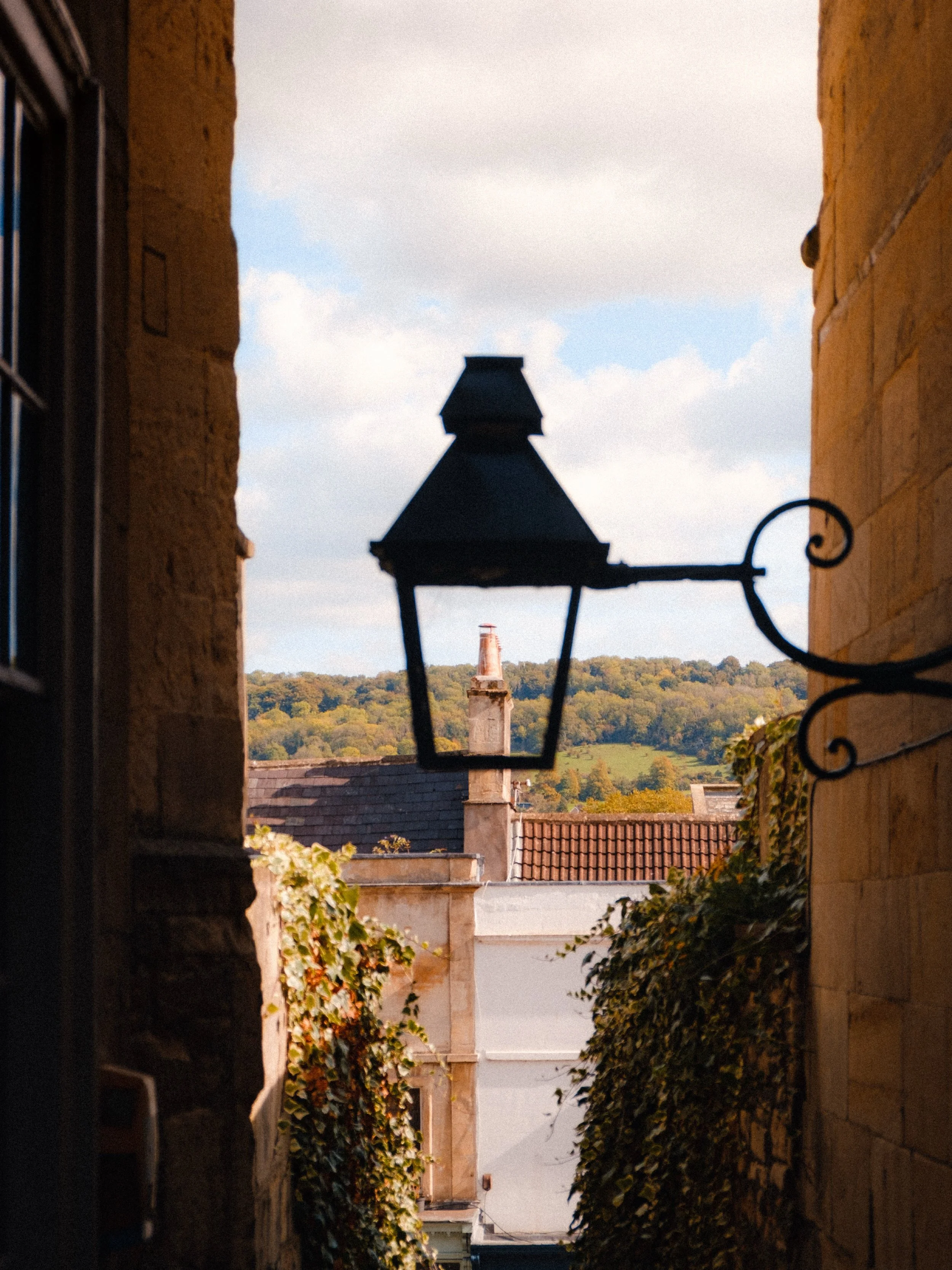 A narrow alleyway between stone buildings with an old-fashioned lamppost in the foreground and a distant hill with trees and chimney in the background.