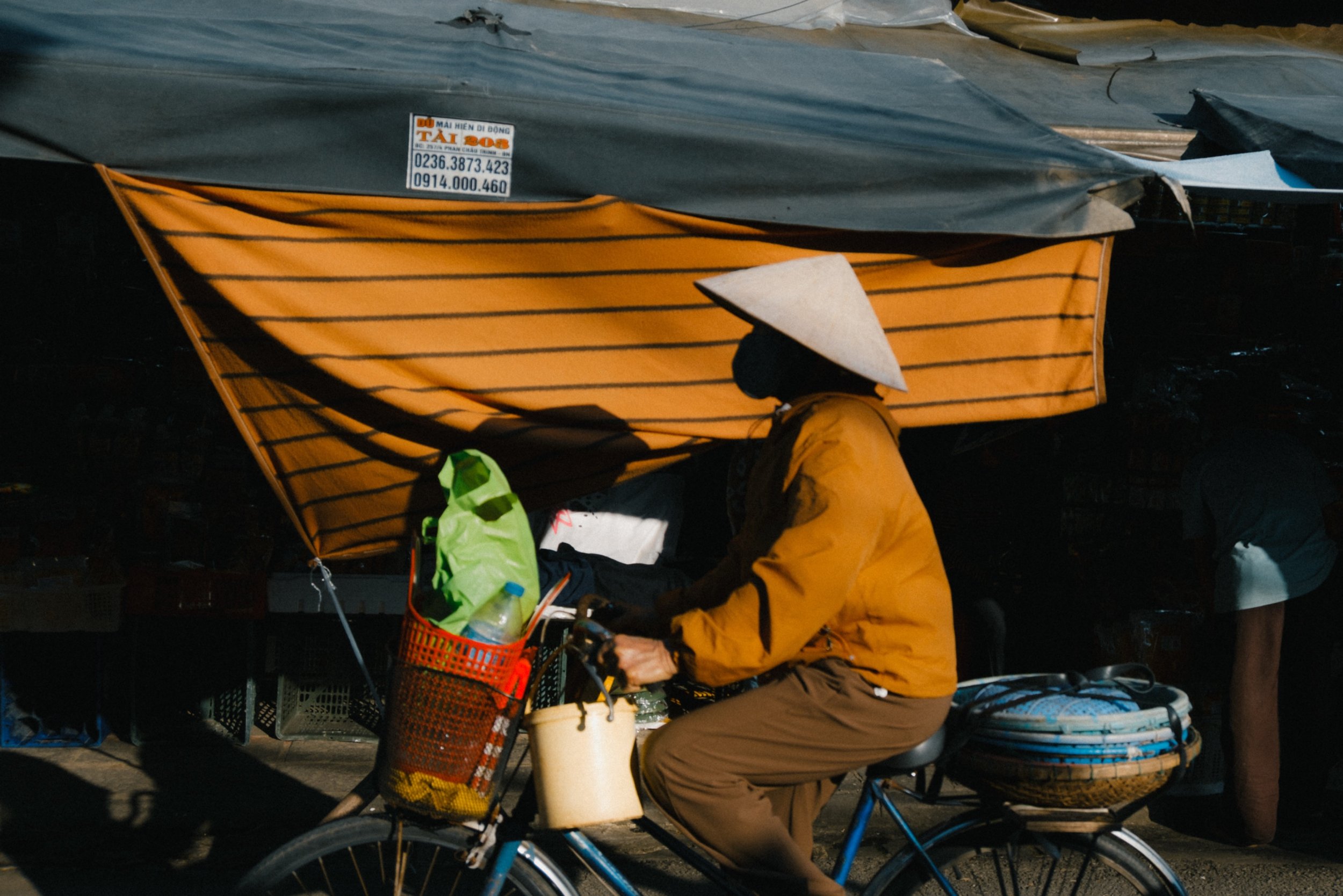 A person riding a bicycle on a street, wearing a conical hat and yellow jacket, with a basket attached to the front of the bike filled with items, under a makeshift shelter or awning.