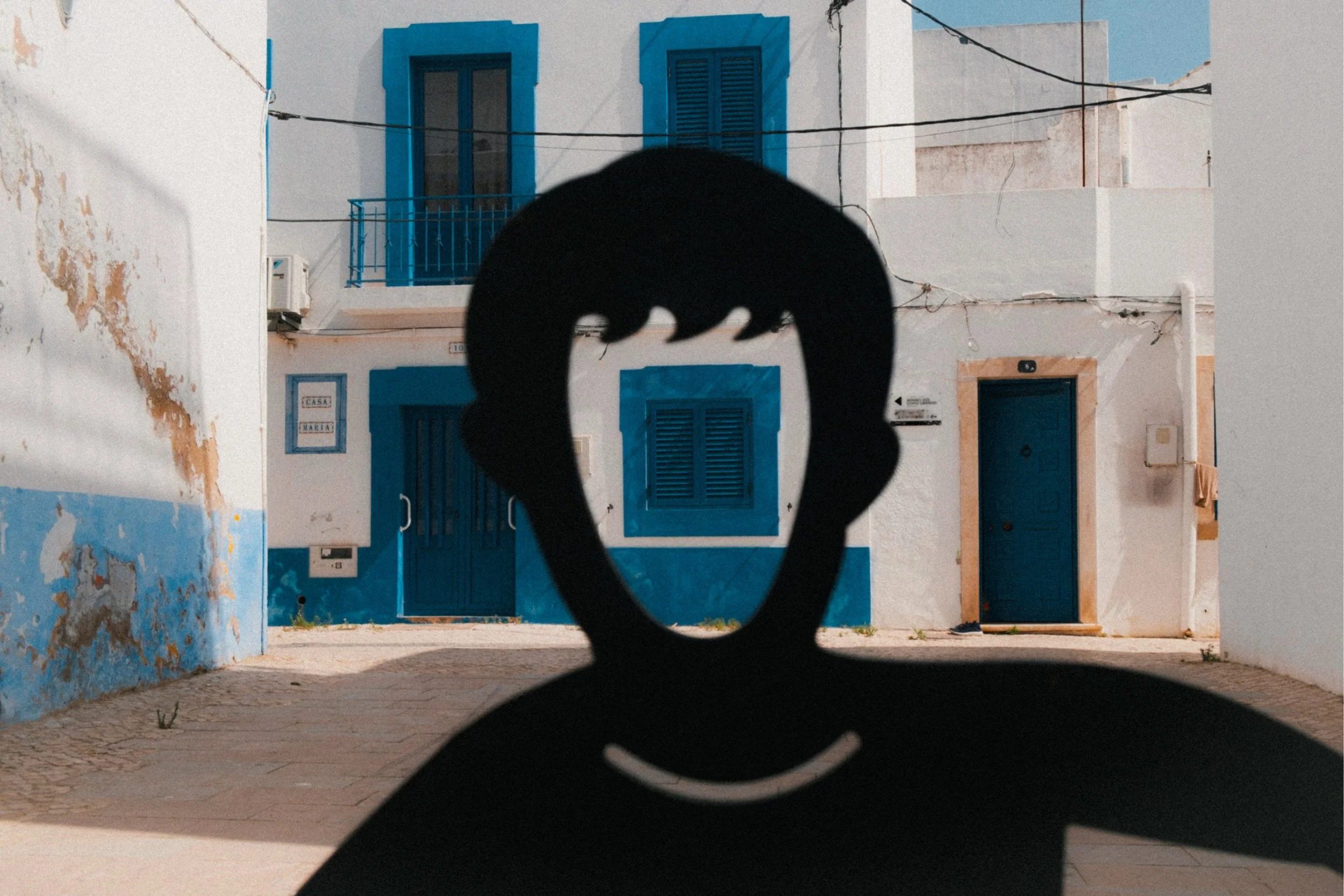 Silhouette of a cat's tail and hind legs in the foreground with white and blue buildings in the background, including closed shutters and a blue door.