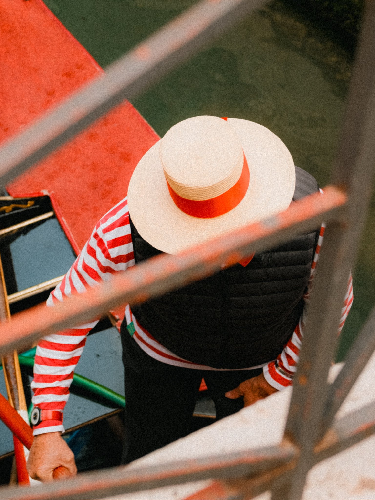 A person wearing a large white sun hat with a red band, a black vest, and a red and white striped shirt, seen from above through a fence or railing.